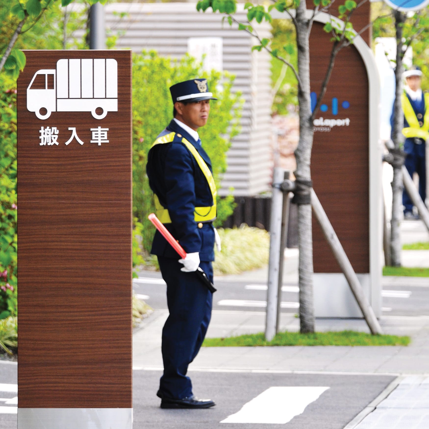 Modern Japanese wayfinding sign with delivery vehicle icon, security staff member, and landscaped entrance at a contemporary facility.