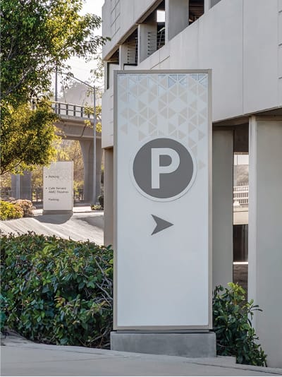 Fashion Valley Mall parking wayfinding pylon featuring a geometric pattern header, bold parking icon, and directional arrow, with a secondary wayfinding sign and parking structure visible in the background.