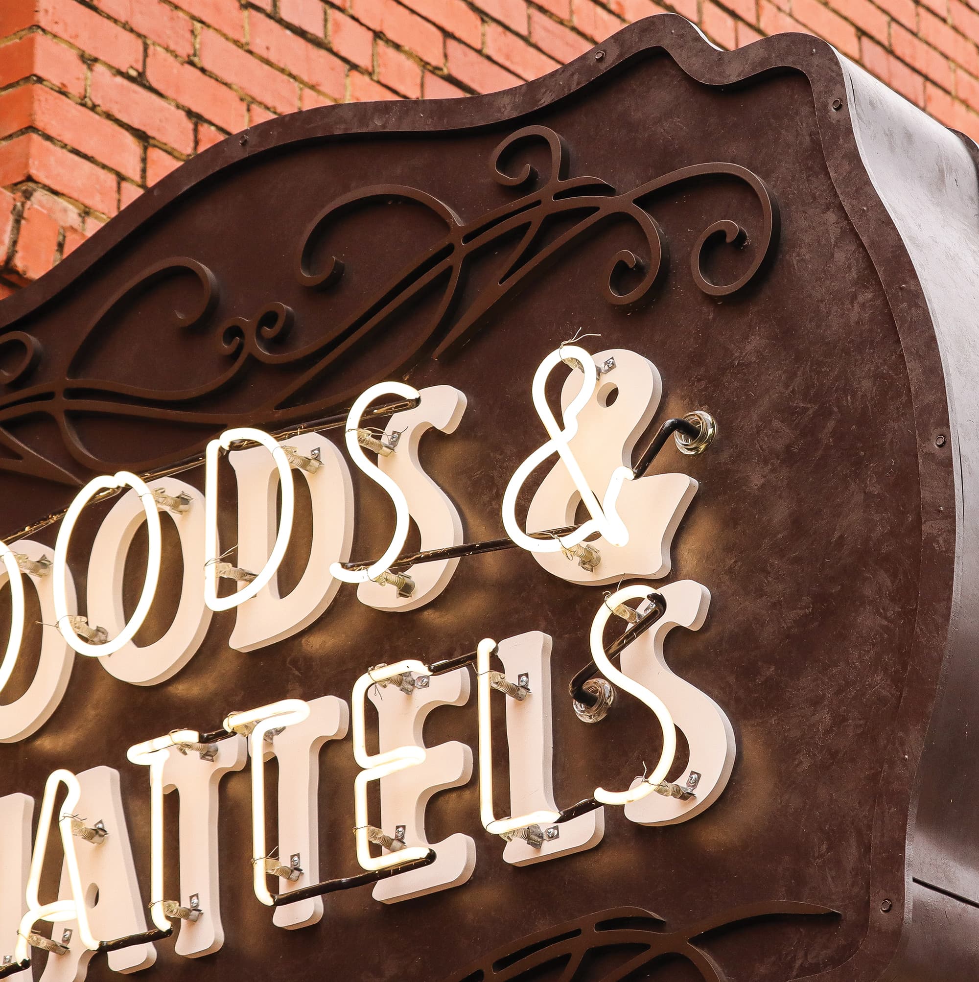Closeup of a sign. Its face is made of weathered, rust-brown metal. The sign’s text, which is cut off in the image so the words can’t be fully read, consists of two layers: behind, a heavy serif font with rounded corners, and in front, a stroke font made of neon light tubes. There is an ornament resembling a calligraphic flourish above and below the lettering, rendered in the rust-brown color of the sign face. A wall is partially visible behind the sign, made of red bricks.
