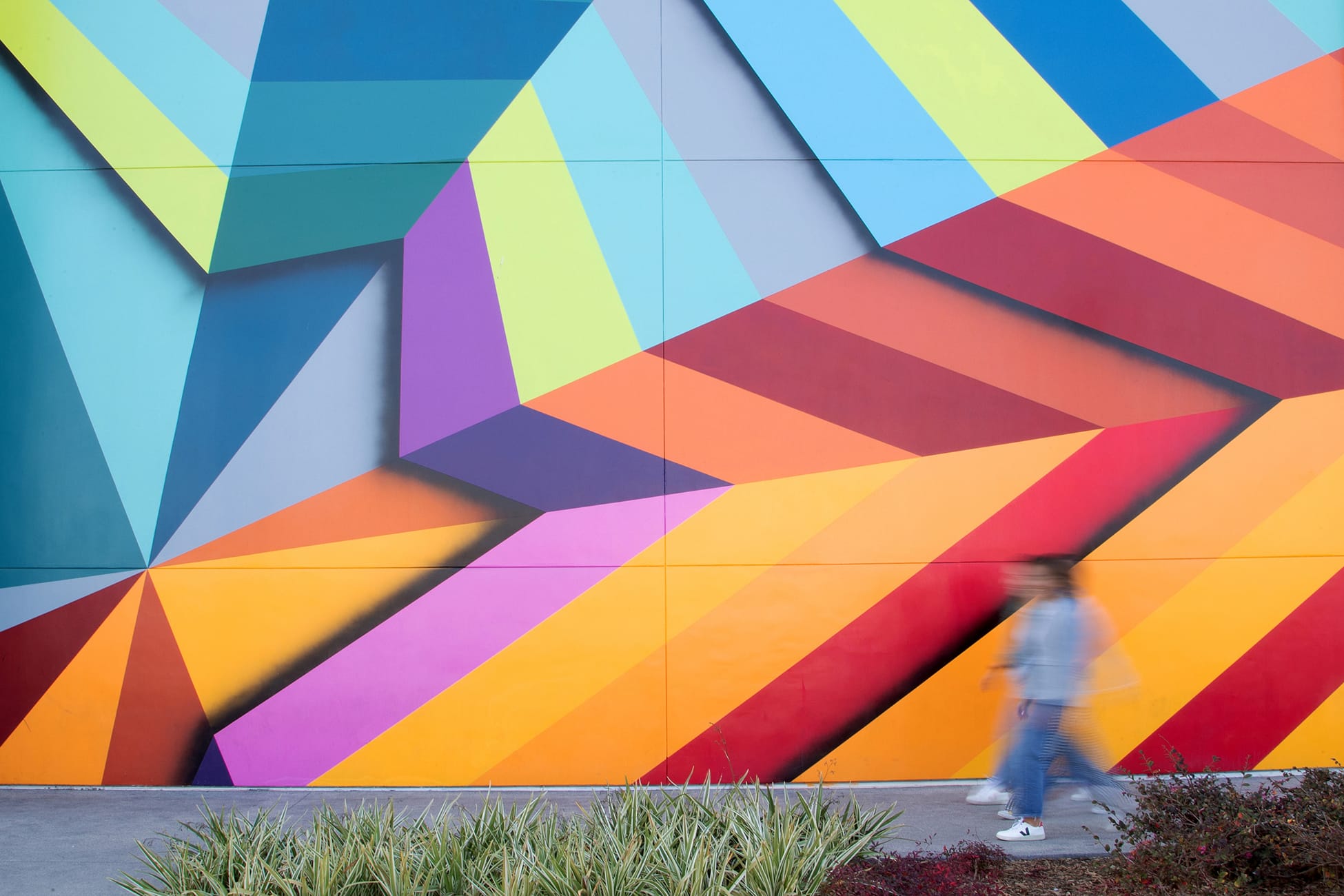 A pedestrian walks past the large-scale geometric mural on the exterior facade of Rodeo 39 Public Market in Stanton, CA, featuring bold interlocking triangular forms in a full spectrum of colors including teal, yellow-green, purple, orange, red, and pink.