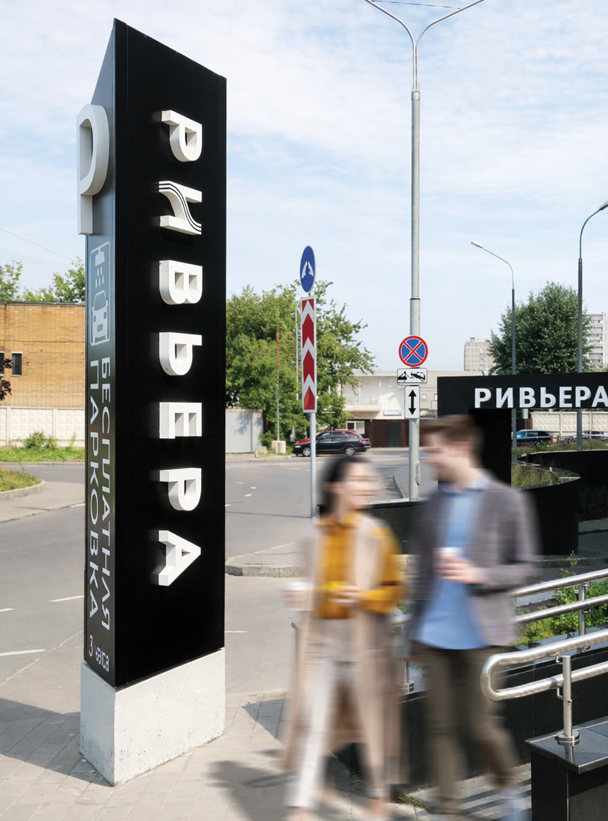 Large-scale exterior Riviera branded pylon monument sign with dimensional Cyrillic letterforms and parking information on an urban streetscape with pedestrians in motion — RSM Design wayfinding and signage design, master sign programs, environmental graphic design, and experiential placemaking for retail, mixed-use, international, and streetscape destinations.