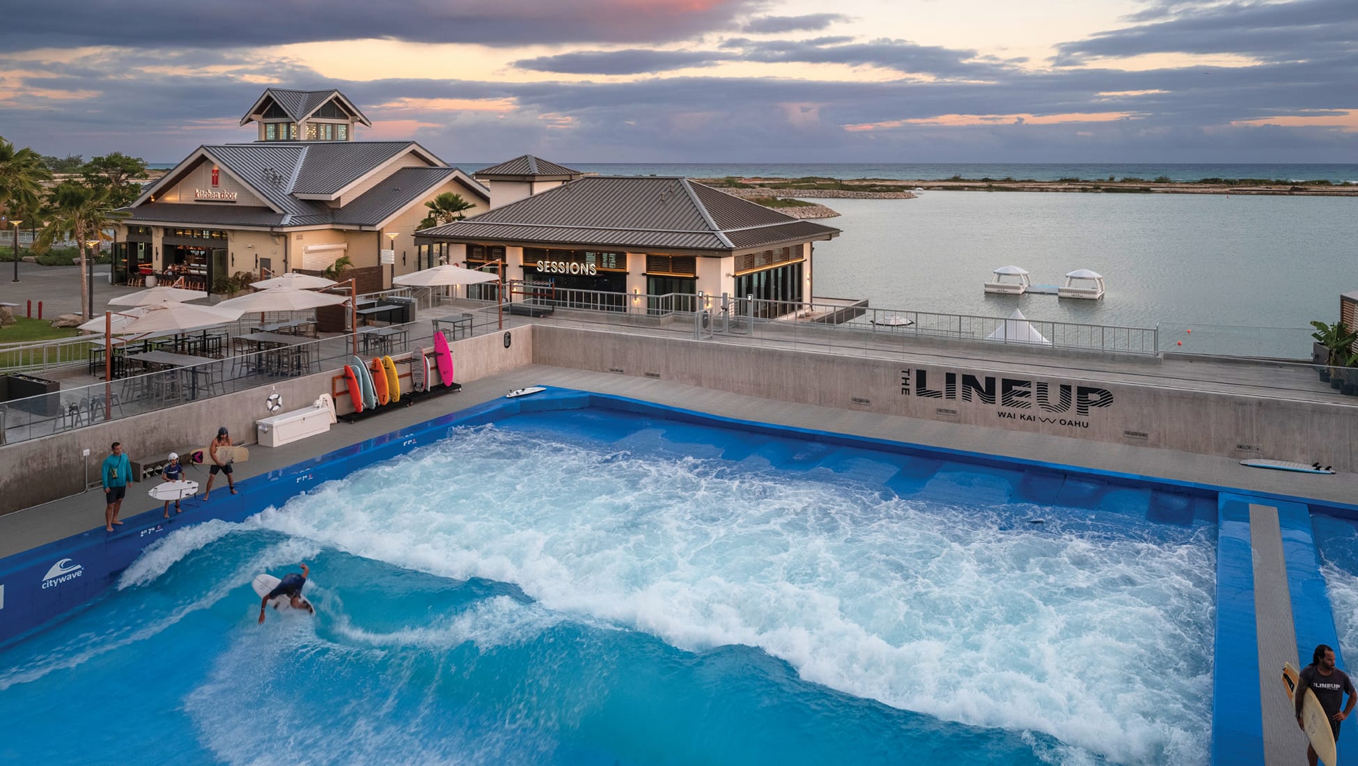 Surfer riding wave in "The Lineup" wave pool at Wai Kai with Sessions and Kitchen Door restaurants and lagoon at sunset, Ewa Beach, Oahu, environmental graphic design by RSM Design.