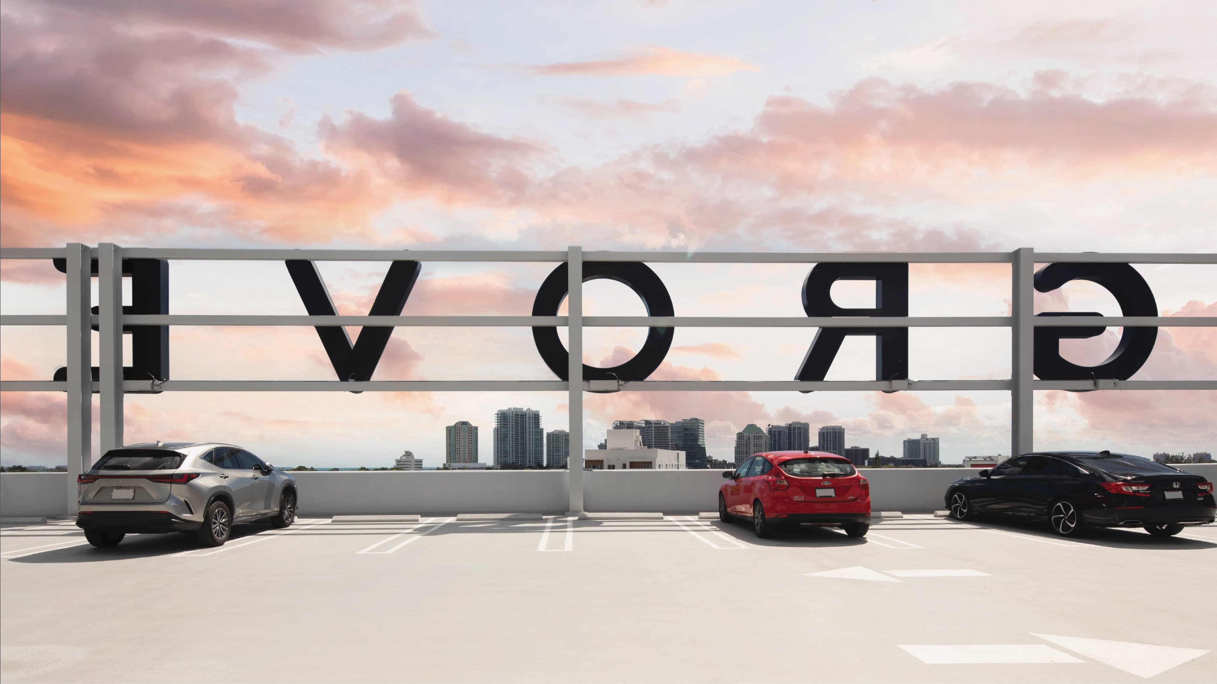 Rooftop parking level with three cars parked beneath a railing displaying large mirrored letters spelling 'Grove' (seen from behind), overlooking the Miami skyline under a pink and orange sunset sky.