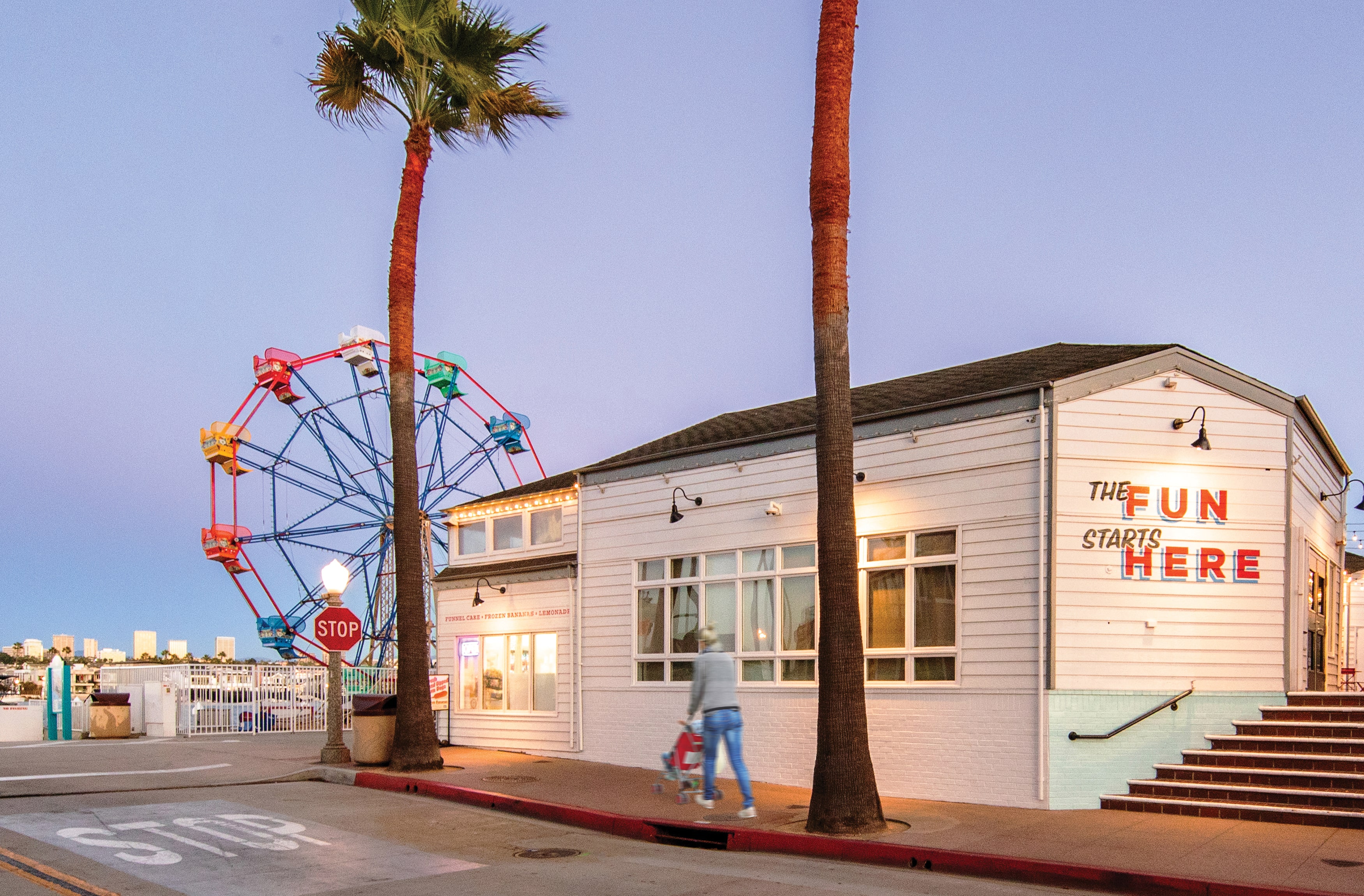 Image of woman pushing a stroller past "The Fun Starts Here" painted red lettering mural on the side of the building at the Balboa Fun Zone in Newport Beach, California nearby the ferris wheel. 