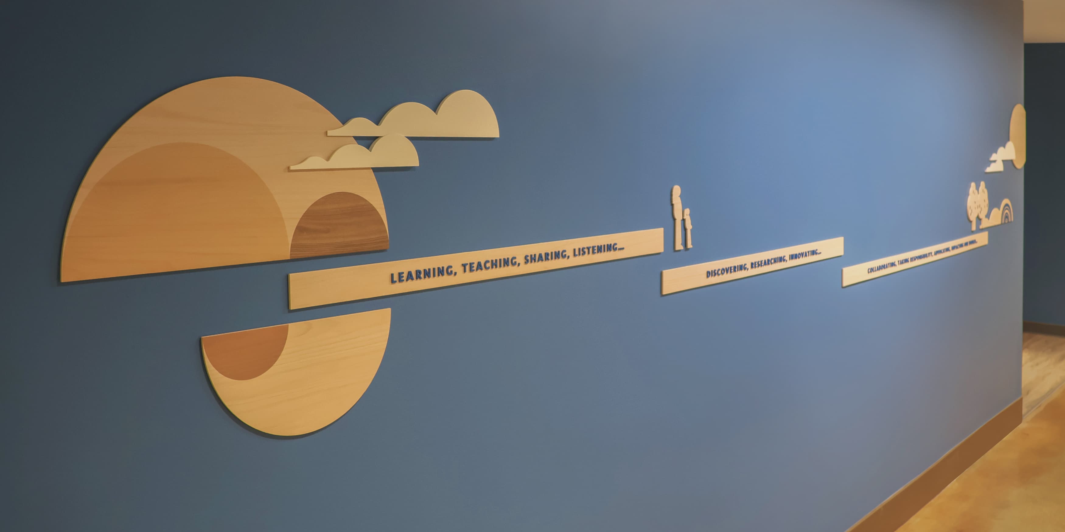 The wall in the image is part of the permanent exhibition designed by RSM Design for the UCLA Fielding School of Public Health's 60th anniversary. It features a vibrant and informative display with a variety of graphic elements and icons. Key public health themes such as Inclusion, Equity, Evidence, Prevention, Community, and Impact are highlighted on the wall, each with its own distinct section. The design includes detailed illustrations, representing different aspects of public health and the school's contributions to the field. The wall is well-lit and incorporates wooden textures with colorful visual elements, making the information engaging and accessible. There are also seating benches integrated into the design, inviting viewers to spend time interacting with the exhibit.