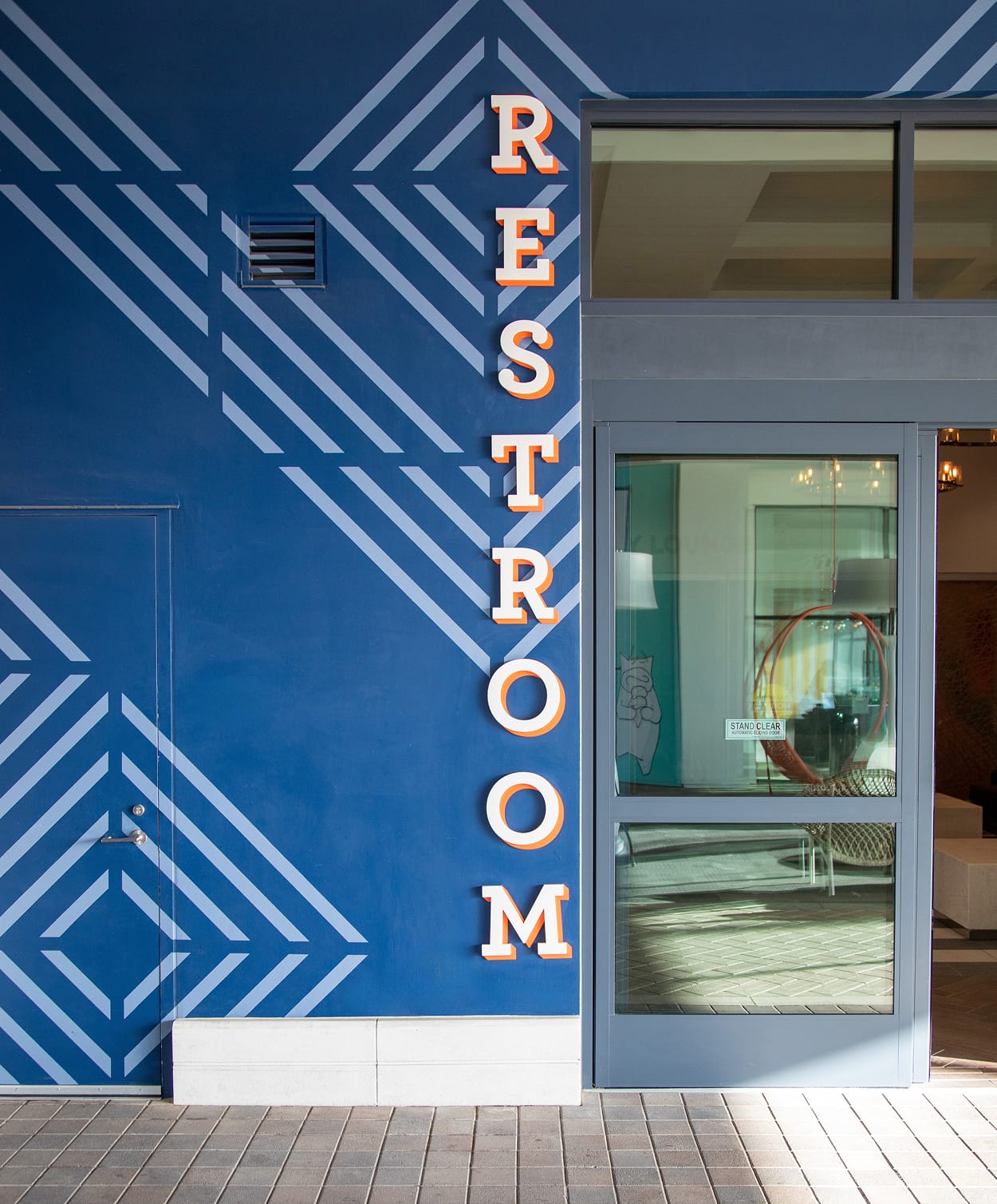 Exterior restroom identification signage at 2nd & PCH in Long Beach, California, featuring vertically stacked dimensional channel letters in white and orange mounted against a bold navy blue geometric patterned wall graphic — an example of RSM Design's wayfinding signage design, architectural graphics, and experiential graphics seamlessly integrated into a coastal waterfront retail and mixed-use destination.