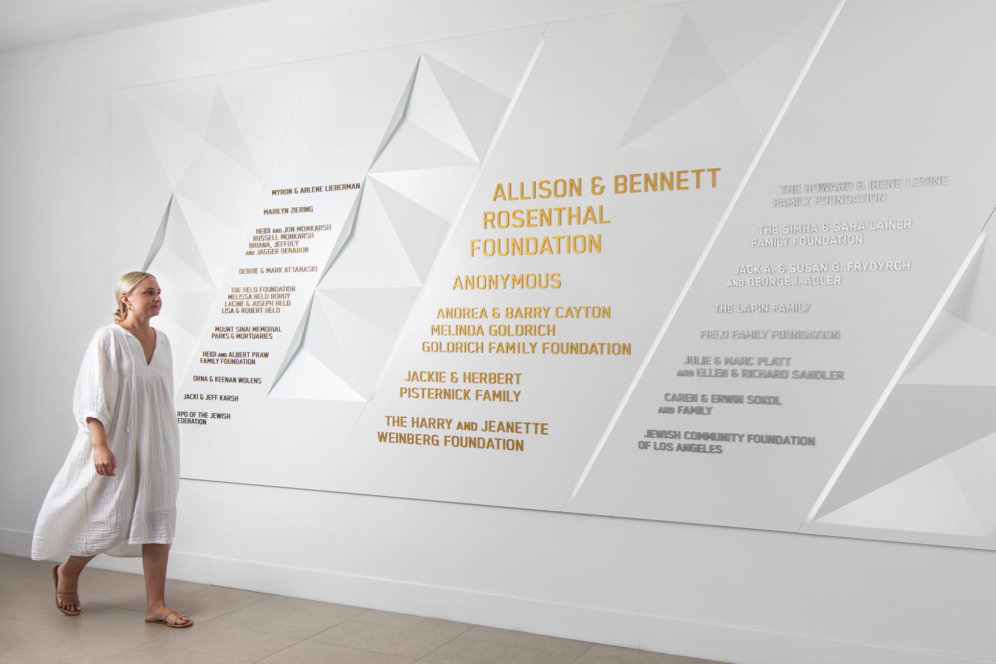Interior donor wall display with three-dimensional geometric white triangular relief panels listing major philanthropic contributors including the Allison & Bennett Rosenthal Foundation, Goldrich Family Foundation, Harry and Jeanette Weinberg Foundation, and Jewish Community Foundation of Los Angeles, with a woman in a white dress viewing the installation