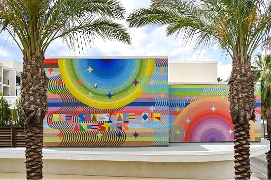 Large-scale exterior mural on the Thompson Palm Springs hotel facade, featuring vibrant concentric arcs, bold stripes, and star motifs in a full spectrum of colors, framed by palm trees against a bright sky.