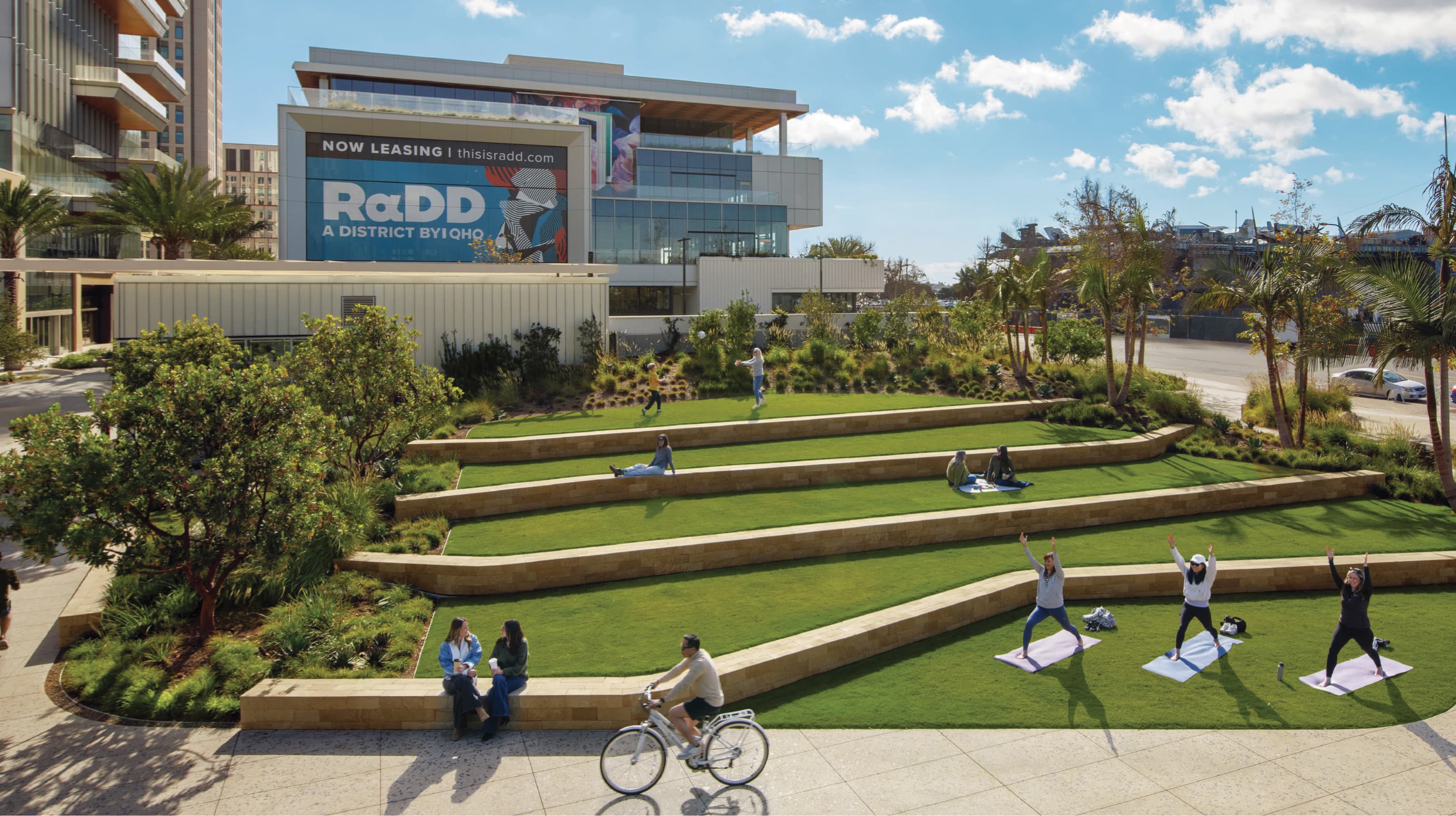 Terraced grass amphitheater at RαDD's waterfront district with people practicing yoga, sitting on the tiered lawns, and playing catch, a cyclist and two women chatting in the foreground, and a large 'RαDD, A District by IQHQ, Now Leasing' banner on a building behind.