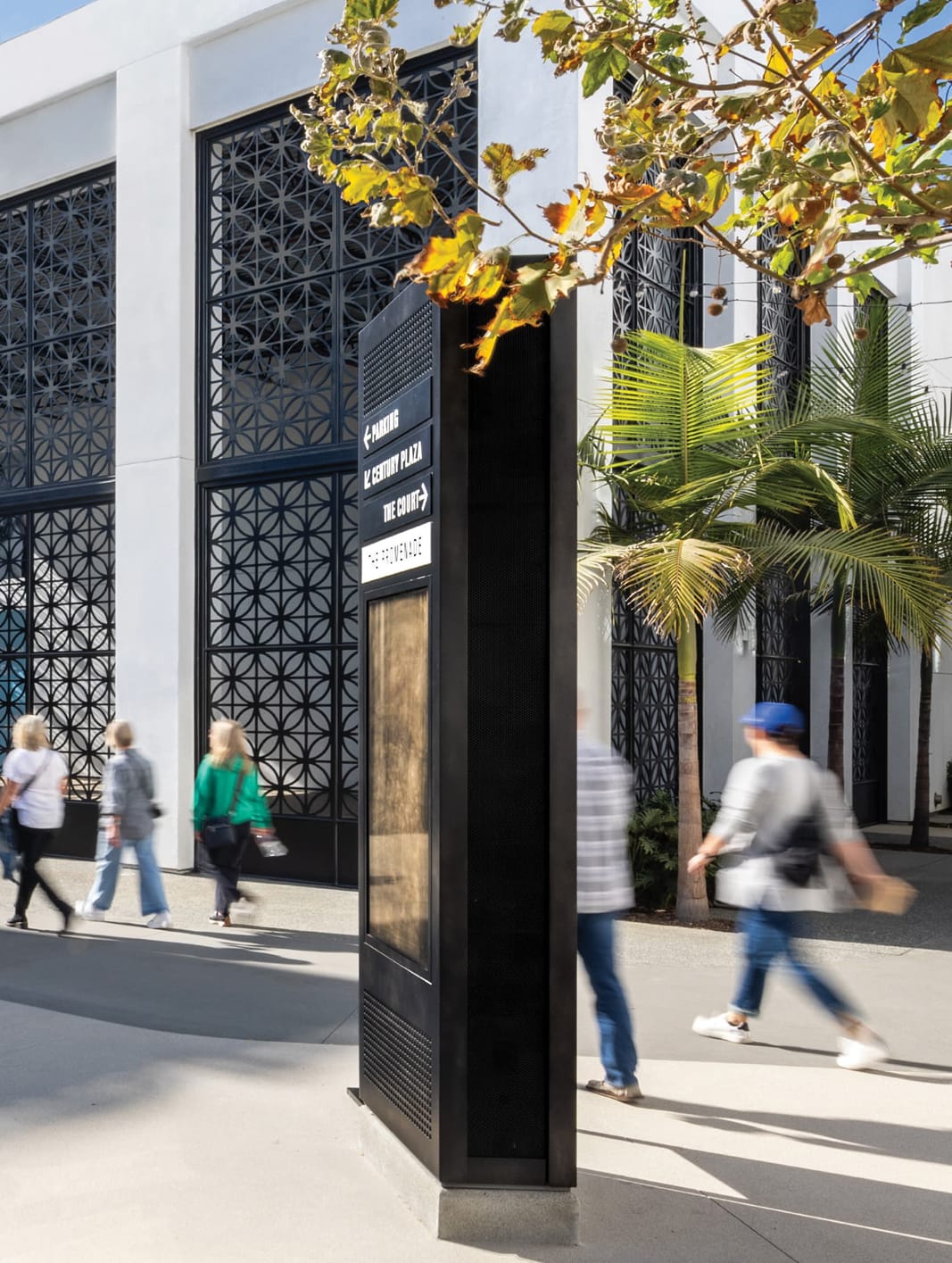 Shoppers walking past Hollywood Park outdoor wayfinding kiosk with directional signs to Parking, Century Plaza, The Court, and The Promenade, with decorative geometric screen facade building in background, Inglewood, California