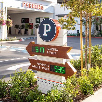 Exterior parking wayfinding pylon sign at 2nd & PCH in Long Beach, California, featuring a branded parking symbol topper with double-sided wood-toned arrow blades displaying live digital LED parking availability counts for Parking & Valet and Parking & Whole Foods — showcasing RSM Design's digital integration and interactive solutions, wayfinding signage design, and master sign program for a coastal waterfront retail, parking garage, and mixed-use destination.