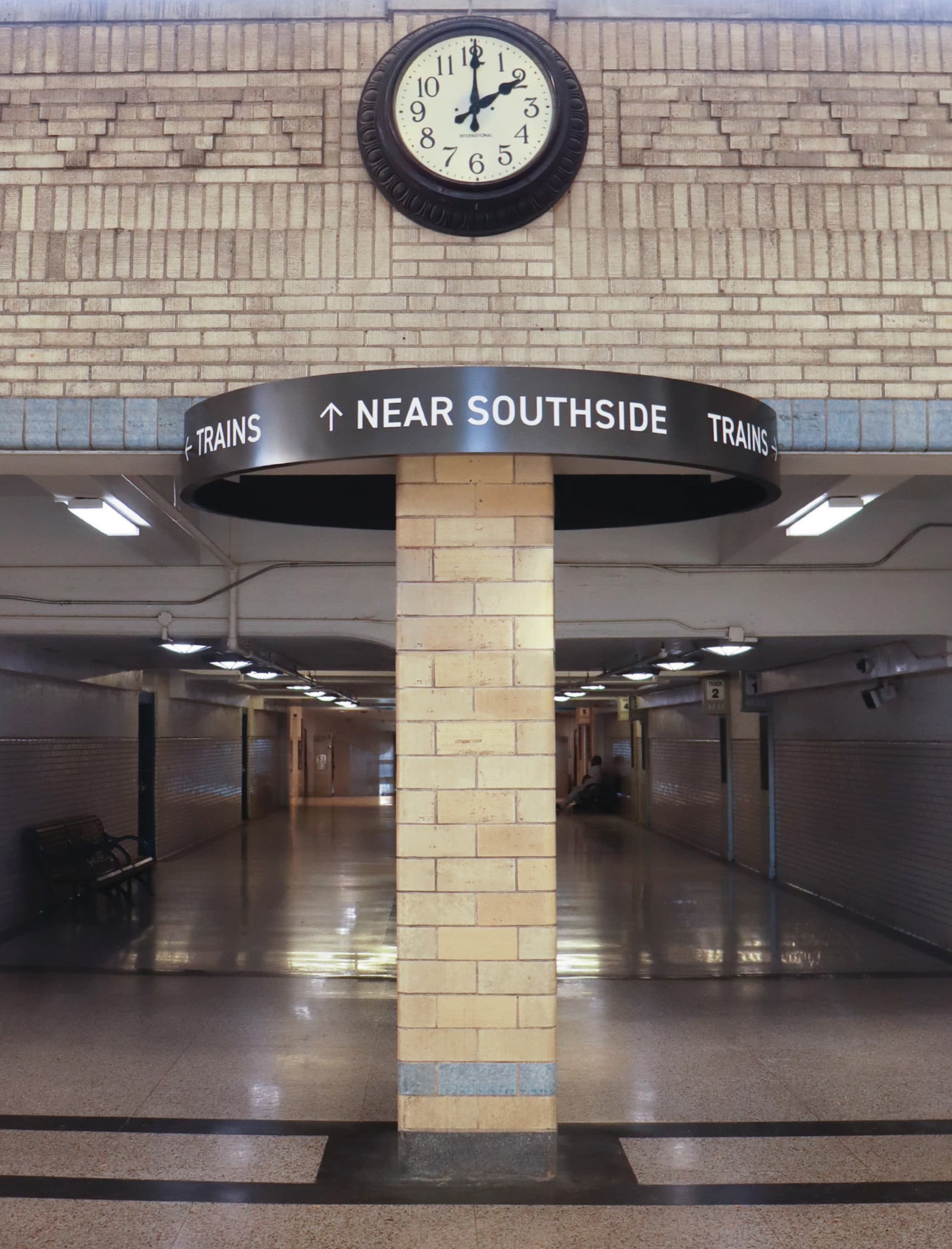 Interior wayfinding ring sign directing passengers to Trains and Near Southside wrapped around a brick column beneath a classic clock in the historic T&P Station concourse, showcasing environmental graphic design and signage for a transit and mixed-use development by RSM Design.