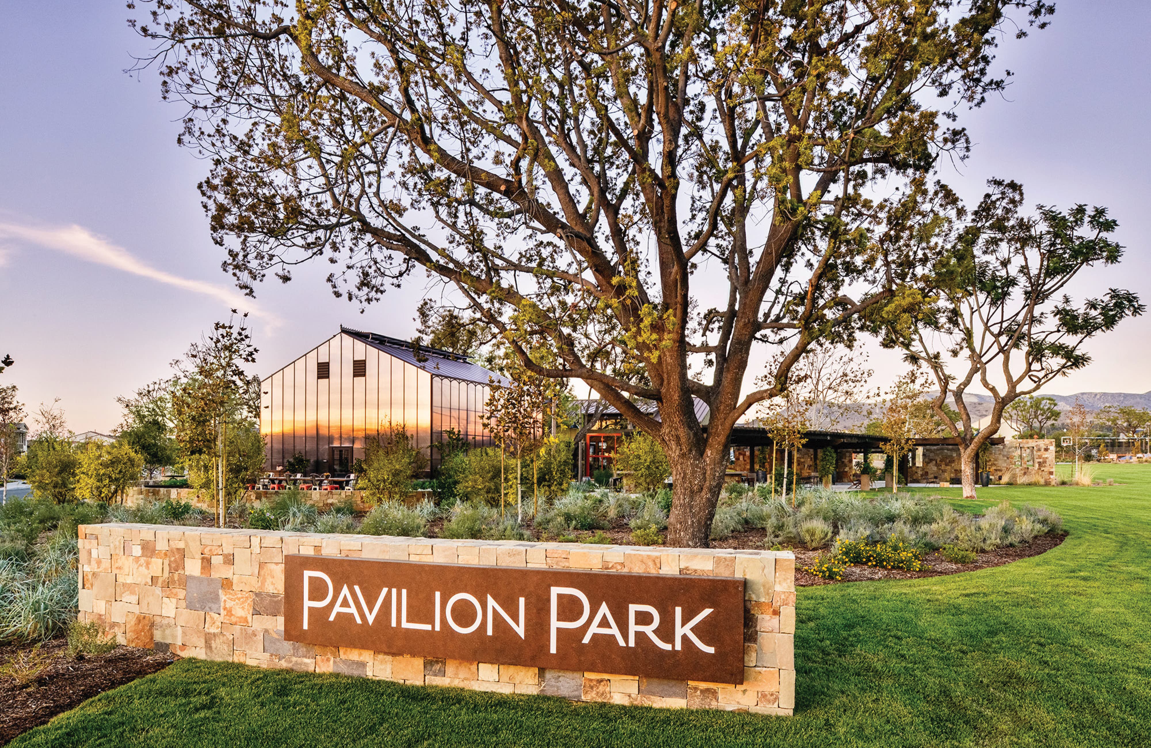 Stone monument sign reading 'Pavilion Park' set in a landscaped lawn, with a mature tree and a glass-walled pavilion building reflecting a colorful sunset in the background, mountains visible in the distance.