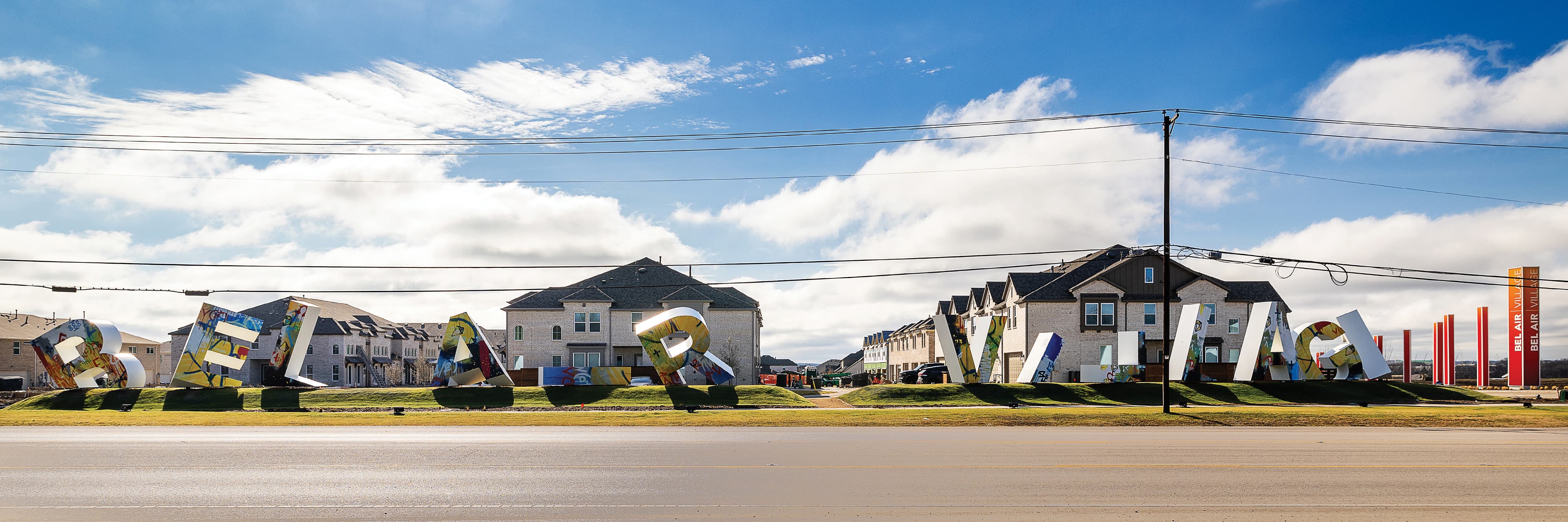 Taken from the street facing a suburban neighborhood of large, brick, 2.5-story, multi-family homes. The letter sculptures stand on a grassy strip between the street and the neighborhood. The illuminated pylons wrap around the corner of the block.