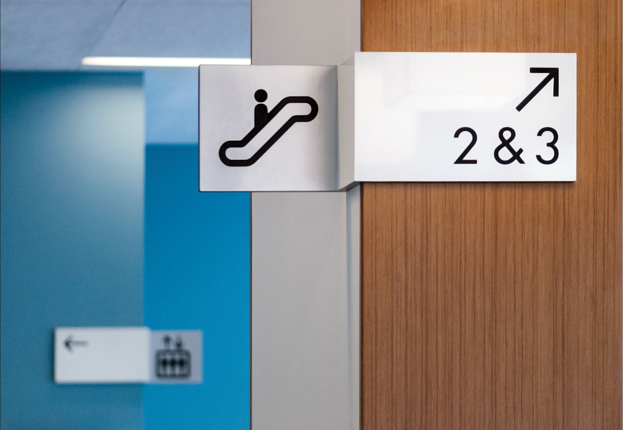 Close-up detail of interior escalator pictogram sign and level 2 and 3 directional wayfinding sign mounted on a warm wood veneer column with brushed aluminum panel, with elevator directional signage visible on a bold blue accent wall in the background at University of the Pacific San Francisco Campus — RSM Design wayfinding and signage design, master sign programs, specialty graphics, and environmental graphic design for education and health and wellness destinations.