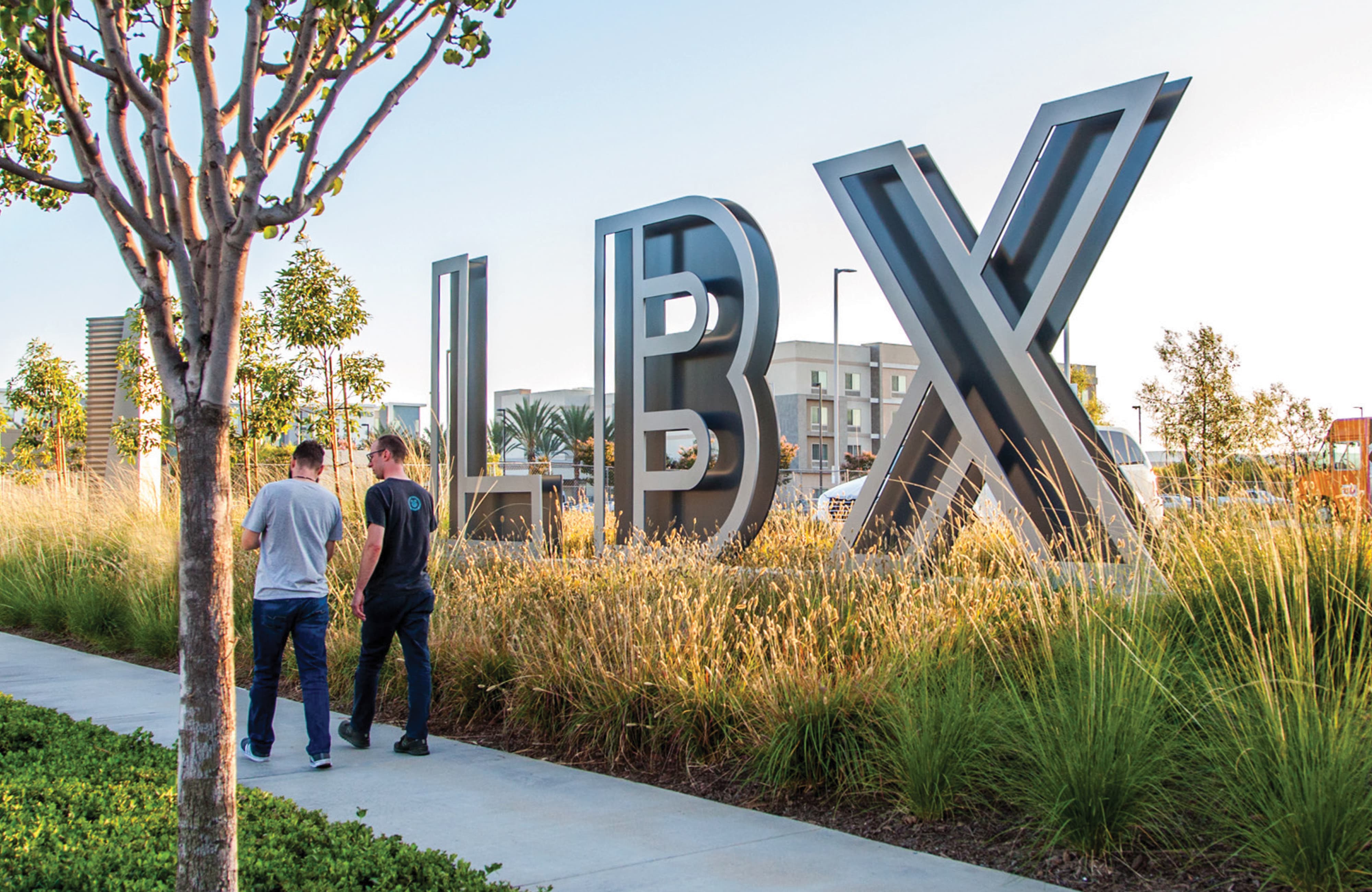 Two men walking along a sidewalk past large dimensional gray 'LBX' letters set among tall ornamental grasses, with buildings and palm trees visible in the background at golden hour.