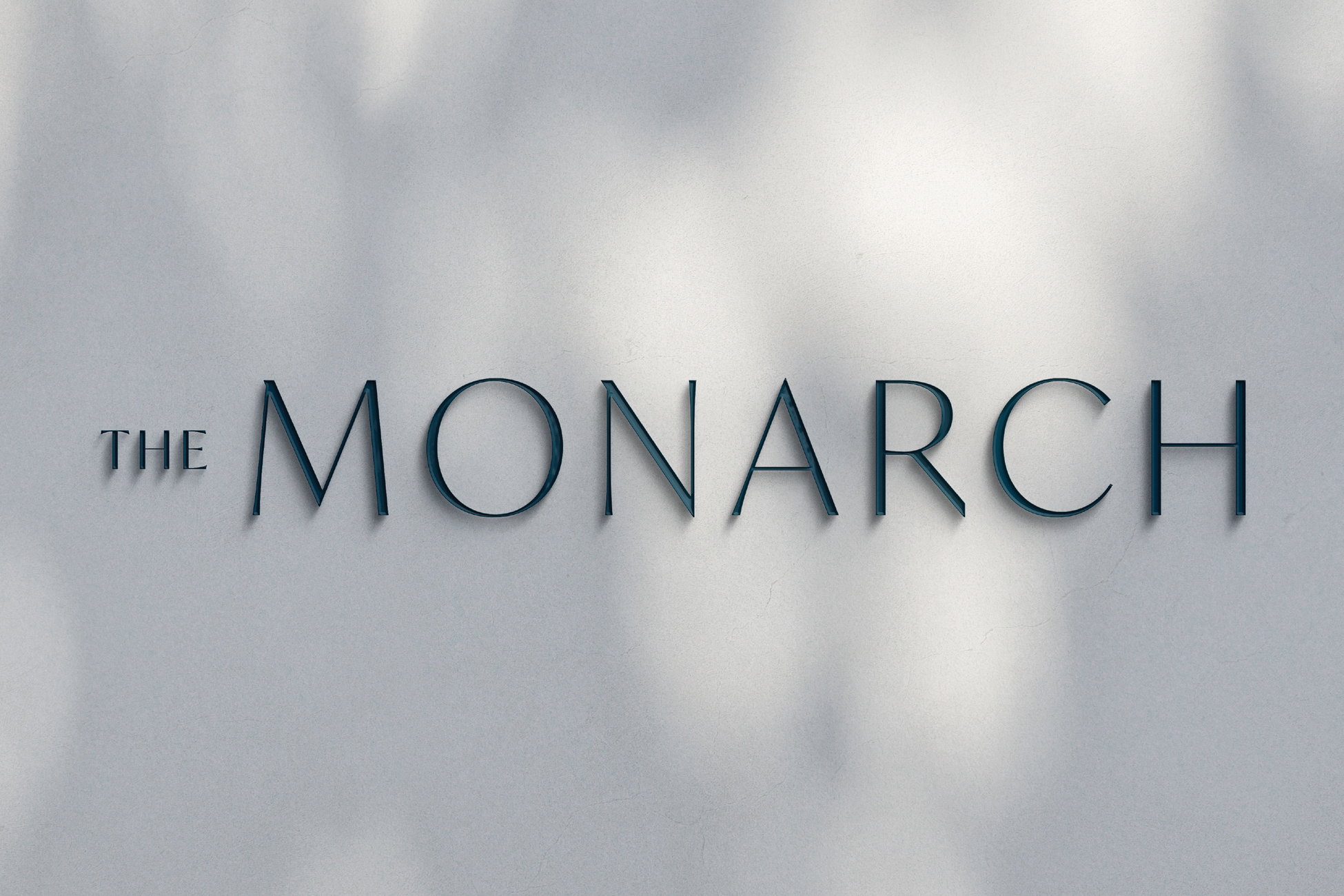 The Monarch at Hall Park dimensional exterior building sign — deep teal fabricated serif letters casting dappled shadow on light grey stucco wall, architectural signage and environmental graphic design by RSM Design