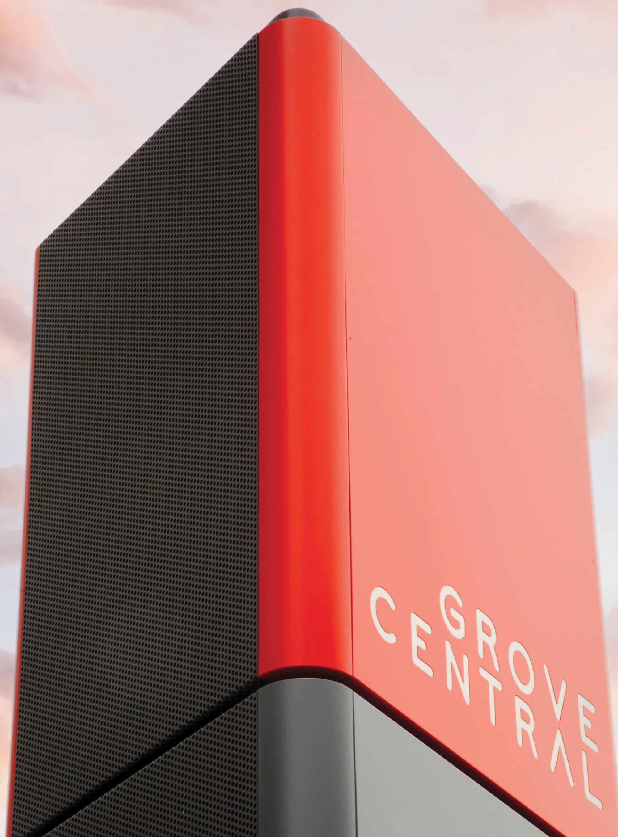Metrorail station platform with a blurred train speeding past, set beside the red and white striped Grove Central parking structure with 'Grove Central' lettering mounted atop the building, birds flying overhead against a partly cloudy sky.