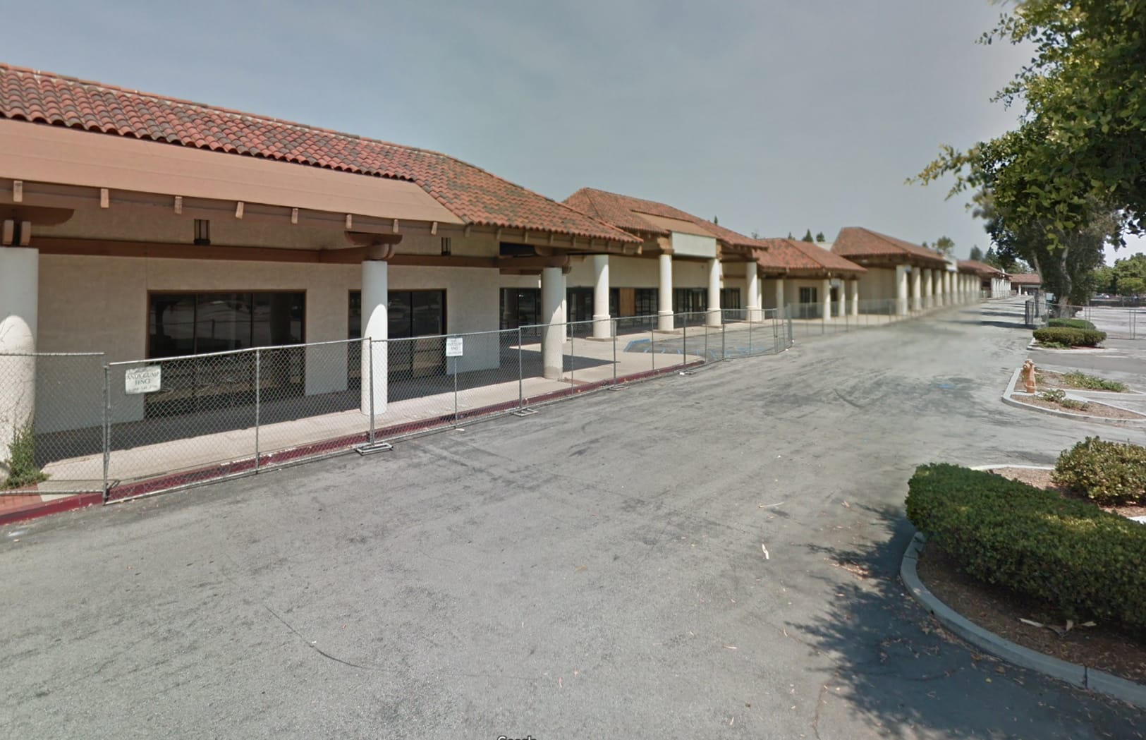 Before photo of the Rodeo 39 project site in Stanton, CA, showing the existing vacant strip mall with dated Spanish-tile roofing, stucco facades, white columns, and chain-link construction fencing along an empty parking lot.