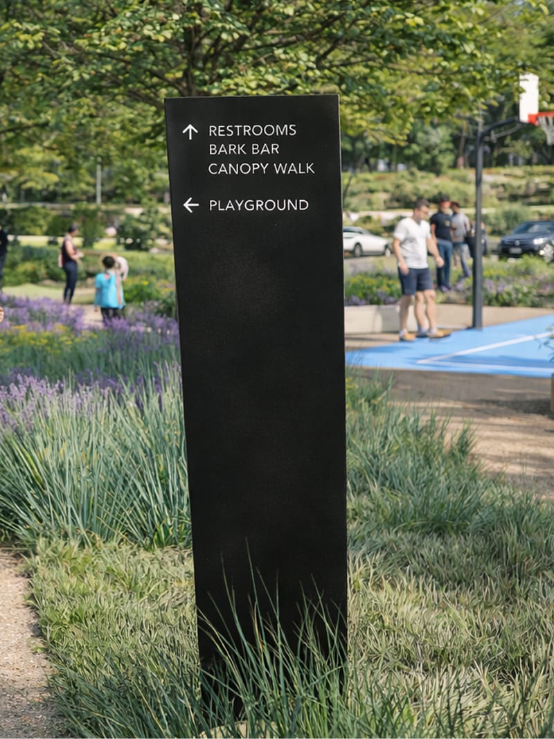 Dark monolithic directional sign in an ornamental grass and lavender garden bed, listing "Restrooms," "Bark Bar," "Canopy Walk," and "Playground" with directional arrows, near a basketball court and gathering park visitors — wayfinding and signage design by RSM Design.
