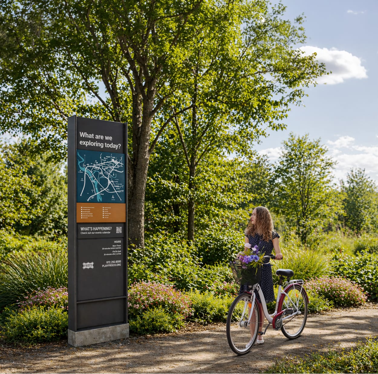 A woman with a bicycle stops to read a dark wayfinding kiosk reading "What are we exploring today?" featuring a trail map and park information, surrounded by lush greenery at Grand Park in Frisco, TX.