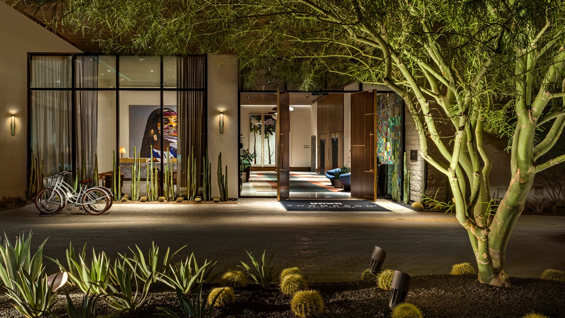 Nighttime exterior entrance of Thompson Hotel Palm Springs, with desert landscaping of columnar cacti, barrel cacti, and agave plants, a palo verde tree in the foreground, and warm lobby lighting visible through open pivot doors and steel-frame windows.
