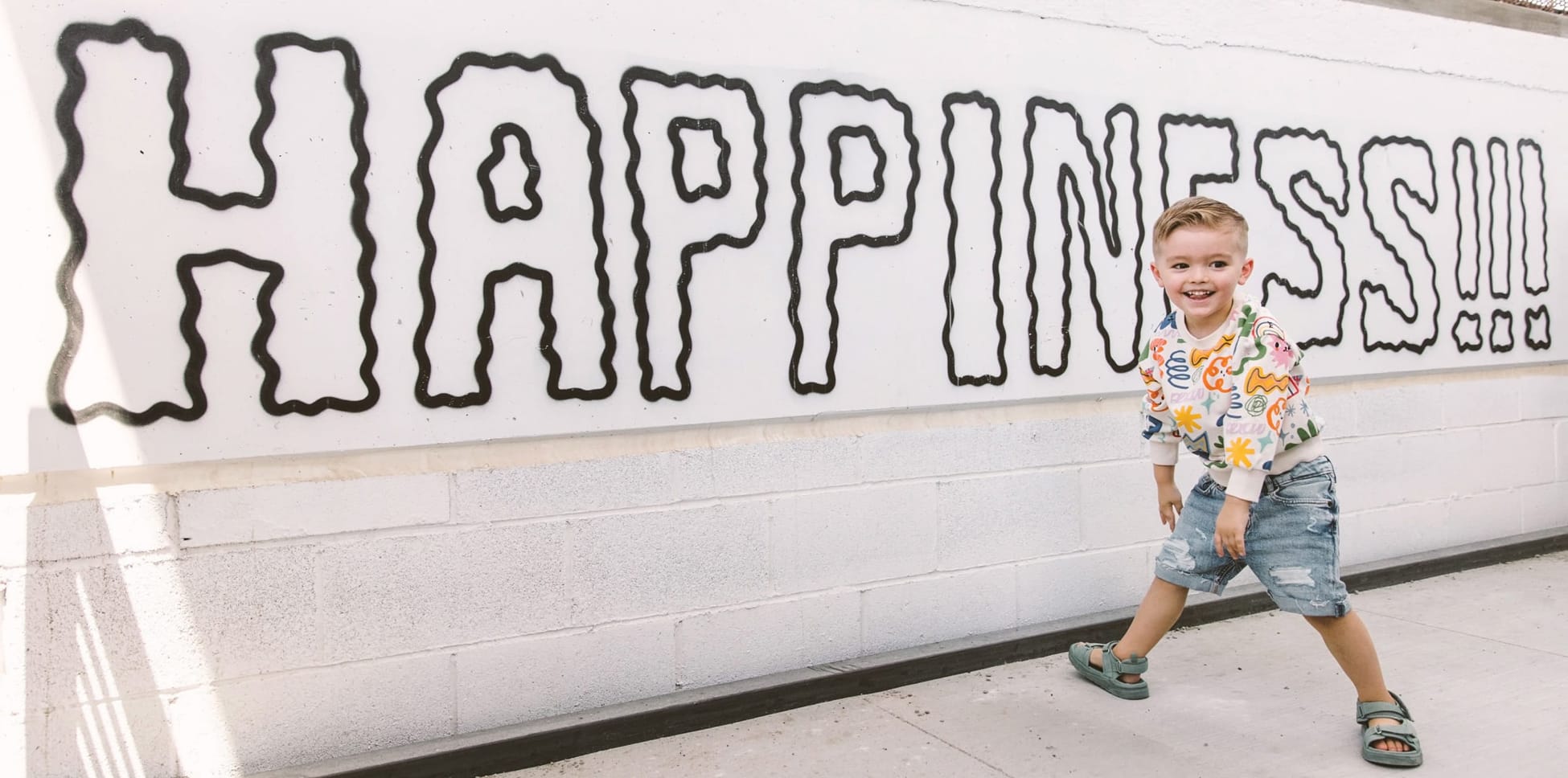 A smiling young child poses in front of a white painted concrete block wall at Uncommons featuring the word "HAPPINESS!!!" in large, bold black outlined bubble letters. The child wears a colorful printed sweatshirt, denim shorts, and sandals.