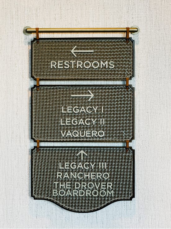 Hotel Drover interior wayfinding sign directing guests to meeting and event spaces including Legacy I, Legacy II, Legacy III, Vaquero, Ranchero, and The Drover Boardroom, with Western-inspired tooled leather texture and brass hardware