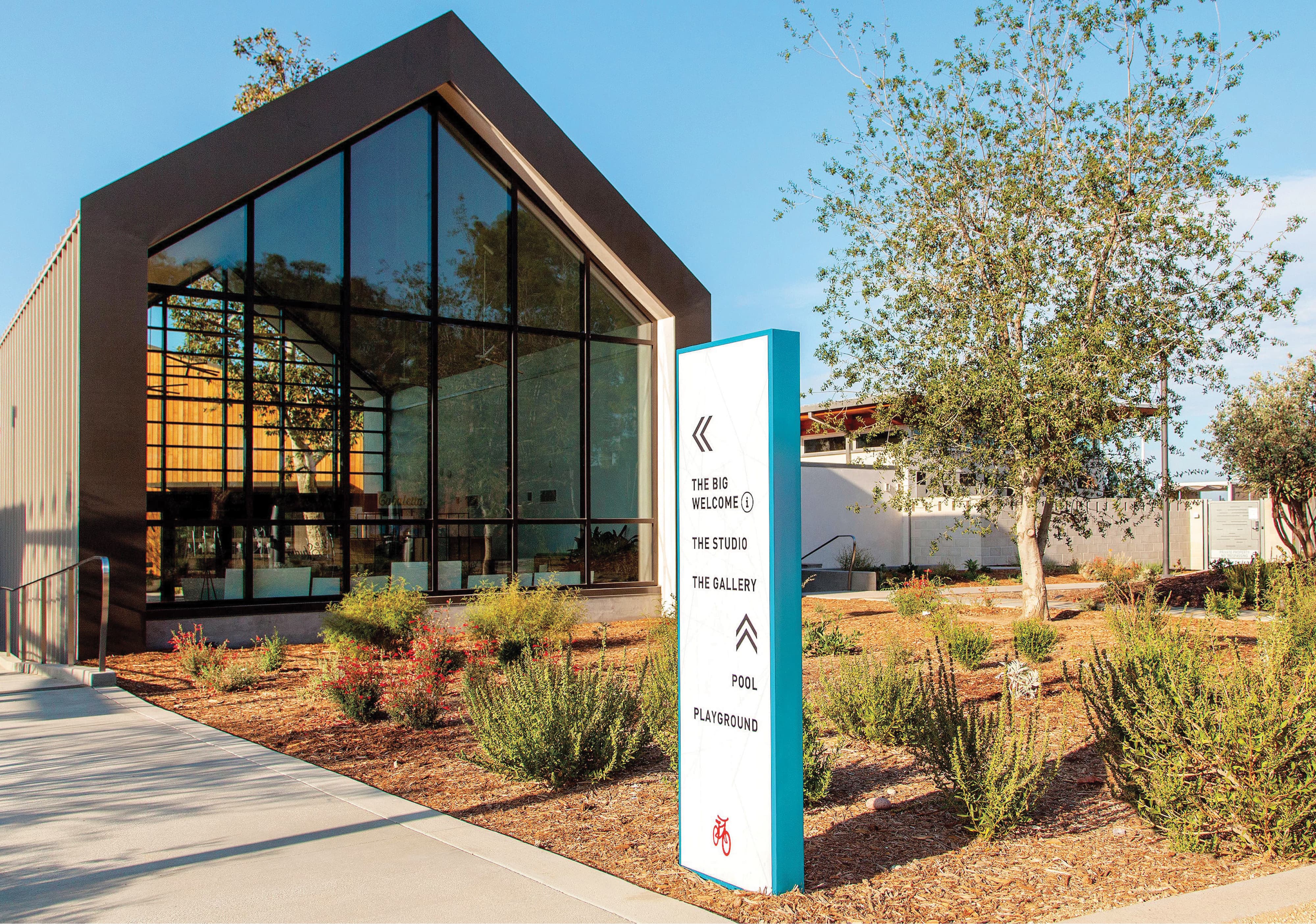 Teal and white outdoor wayfinding totem directing residents to amenities including The Big Welcome, The Studio, The Gallery, Pool, and Playground, set against a modern glass and steel community clubhouse, showcasing placemaking and signage design for a residential community by RSM Design.