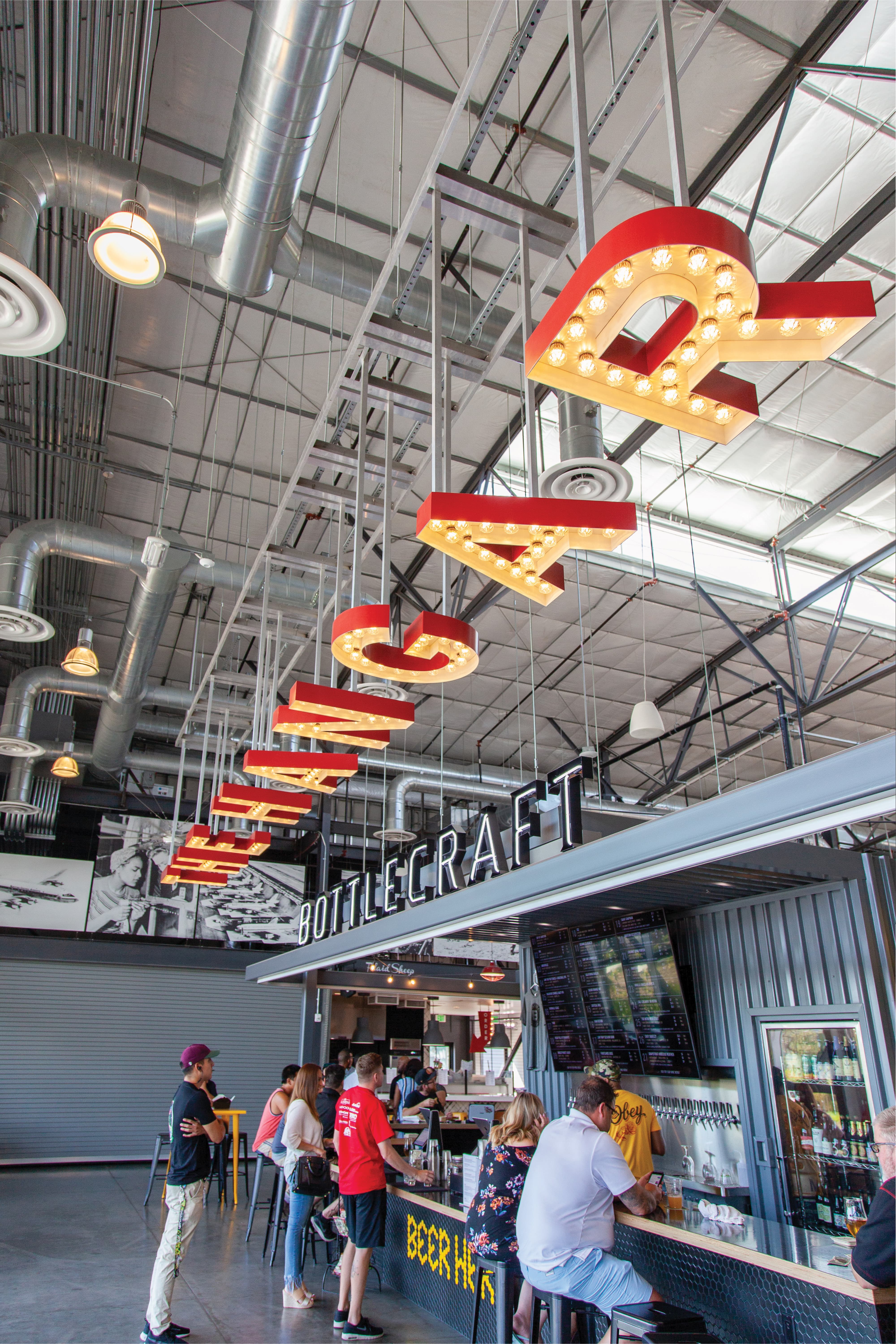 Illuminated marquee letters spelling 'The Hangar' suspended diagonally from an industrial ceiling above the 'Bottlecraft' bar, where customers stand and sit at the counter beneath a taps wall and menu screens.
