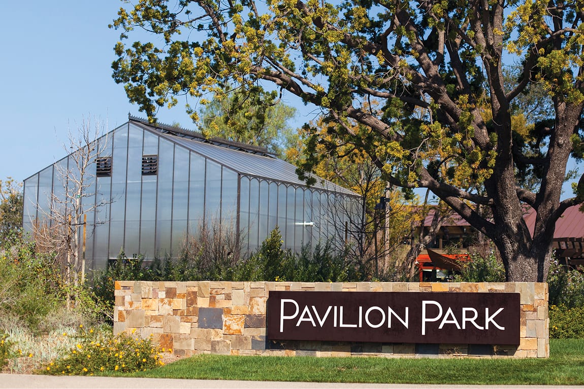 Pavilion Park neighborhood entrance monument sign with white dimensional letters on a dark panel set into a natural stacked stone wall, with a glass greenhouse structure visible in the background in Irvine, California, designed by RSM Design