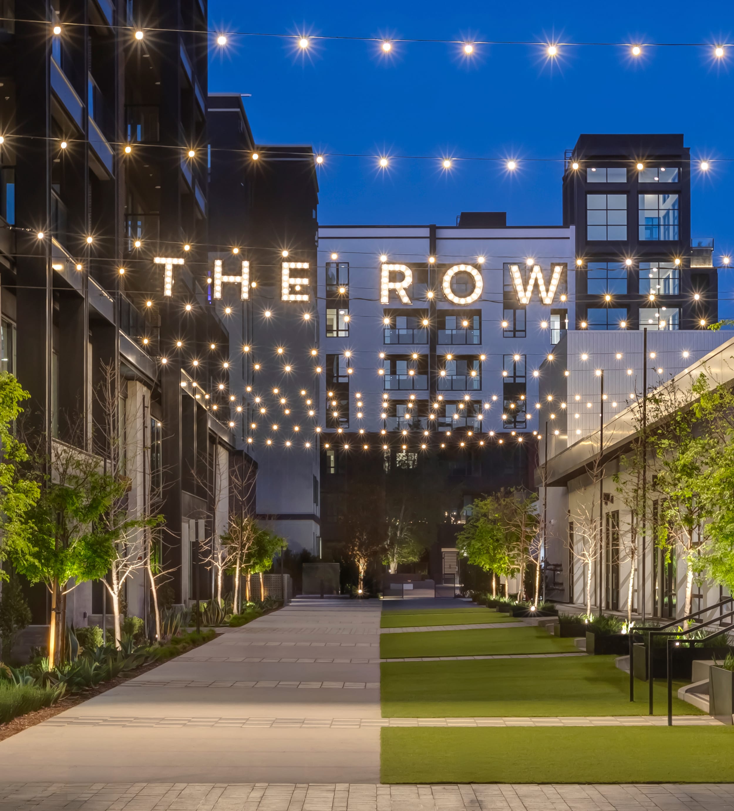 Illuminated marquee-bulb "THE ROW" letters strung above a lawn walkway lined with string lights and trees at twilight, at The Row at Red Hill – placemaking and environmental graphic design by RSM Design.