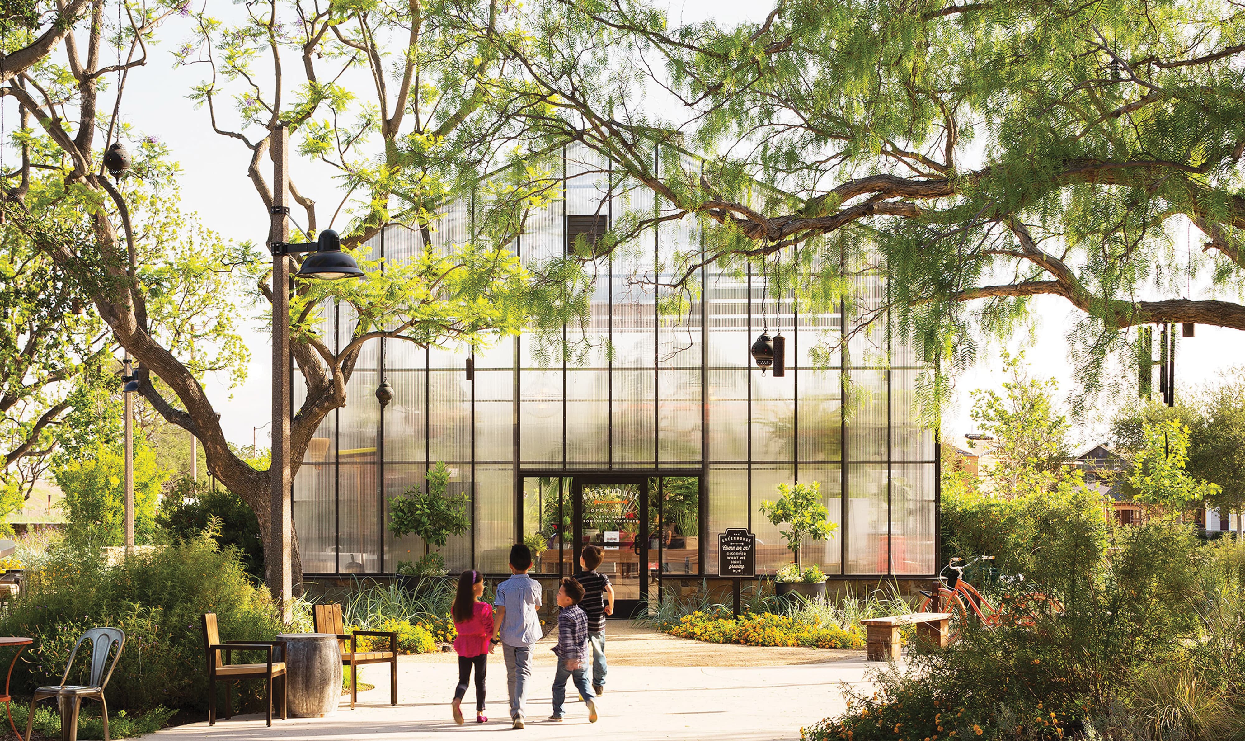 Four children walking toward the entrance of a translucent glass greenhouse building with an 'Open up! Discover what we have growing' chalkboard sign, shaded by large trees with hanging lanterns, garden seating, and flowering shrubs along the path.