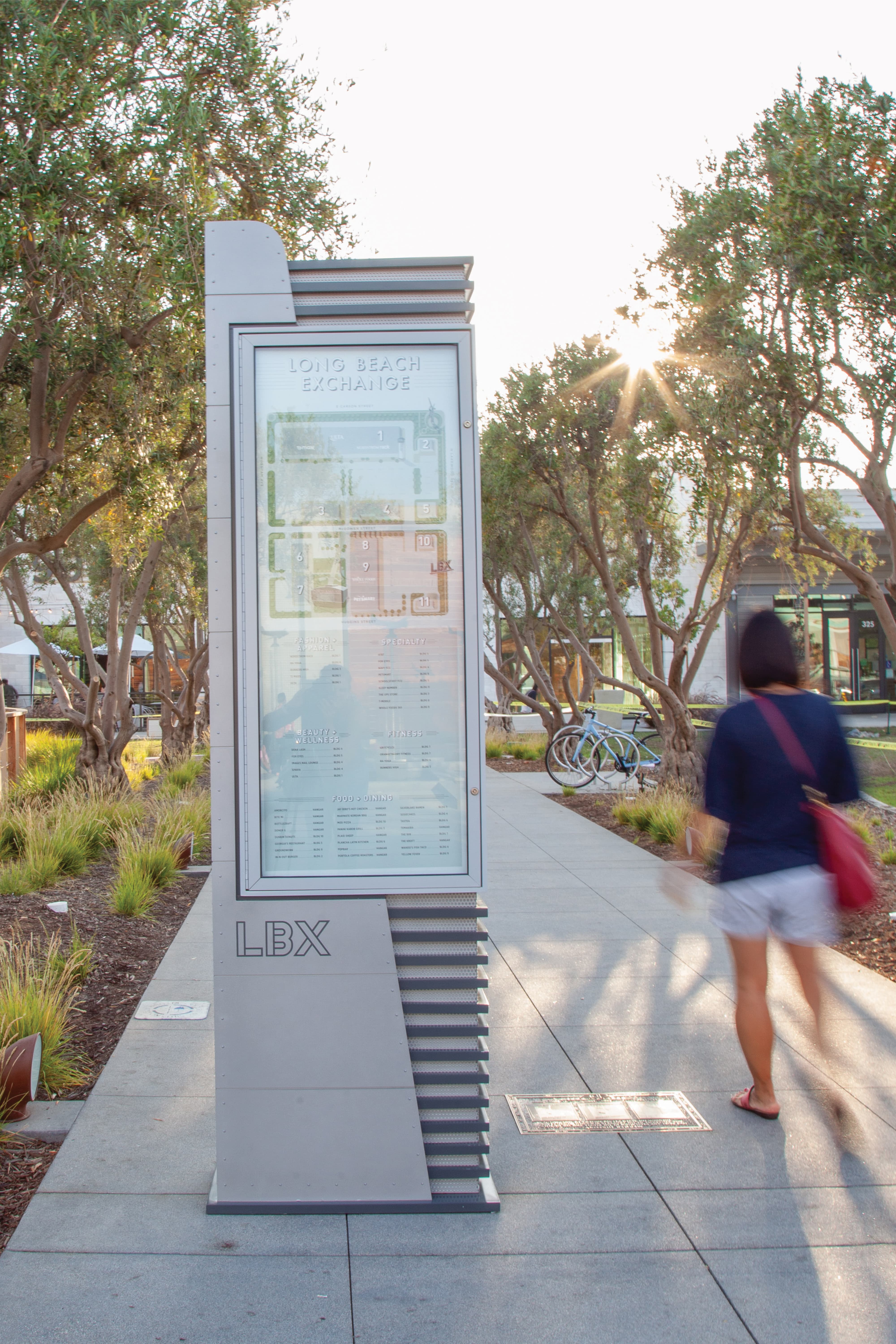 Tall 'LBX' wayfinding sign reading 'Long Beach Exchange' with a color-coded site map and category directory for fashion, beauty, fitness, and dining, set on a tree-lined paved path as a woman walks past bicycles at sunset.