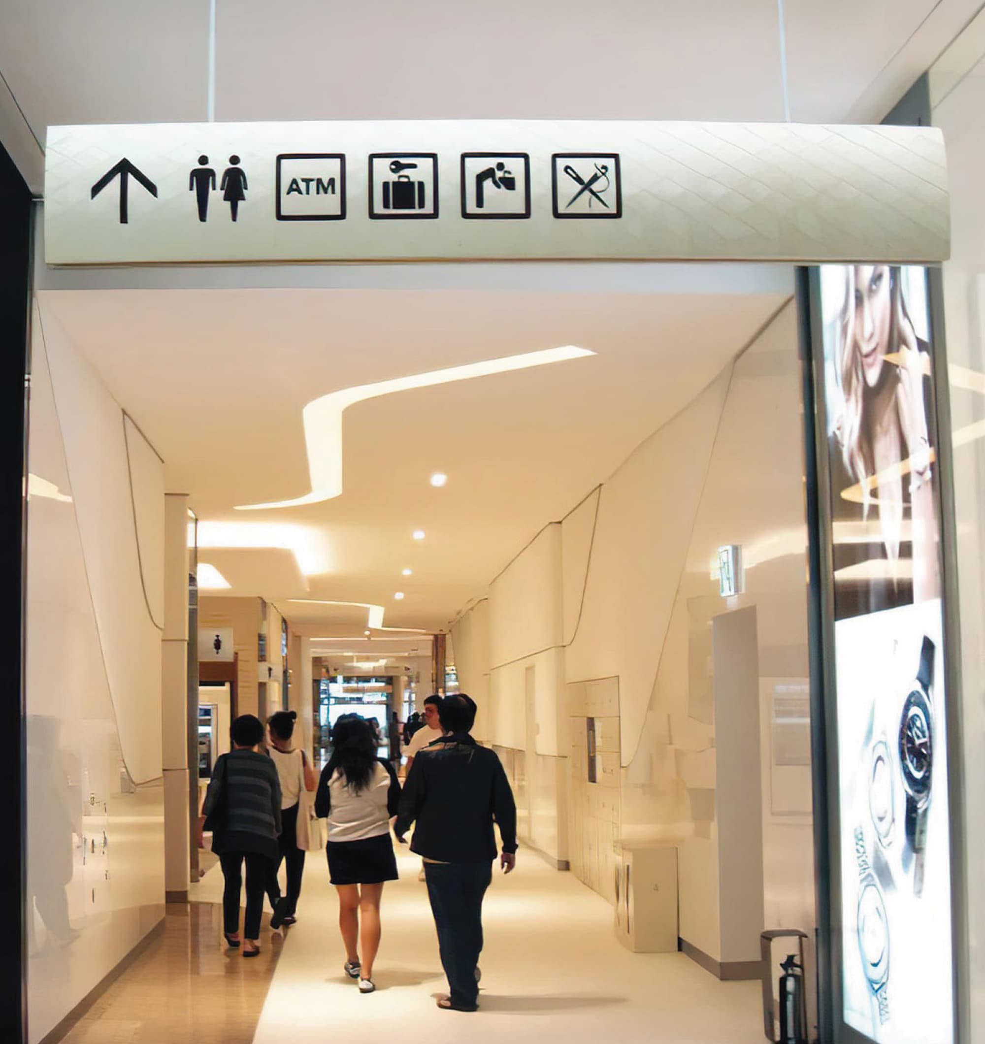 Overhead mall wayfinding sign with icons for restrooms, ATM, luggage storage, and other services above a bright hallway, where shoppers walk past a jewelry storefront and locker area.