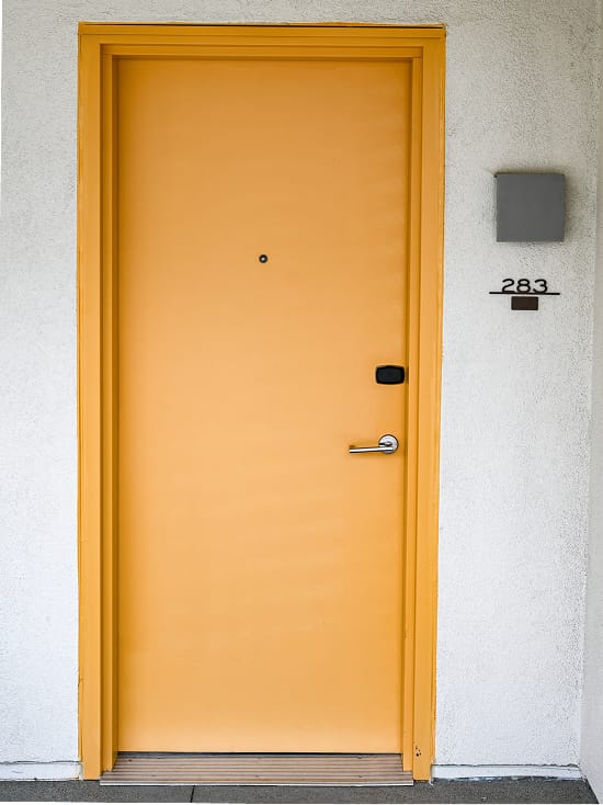 Vibrant yellow-orange guest room door numbered 283 at Thompson Palm Springs, with a chrome lever handle and modern room number placard against a white stucco exterior wall.