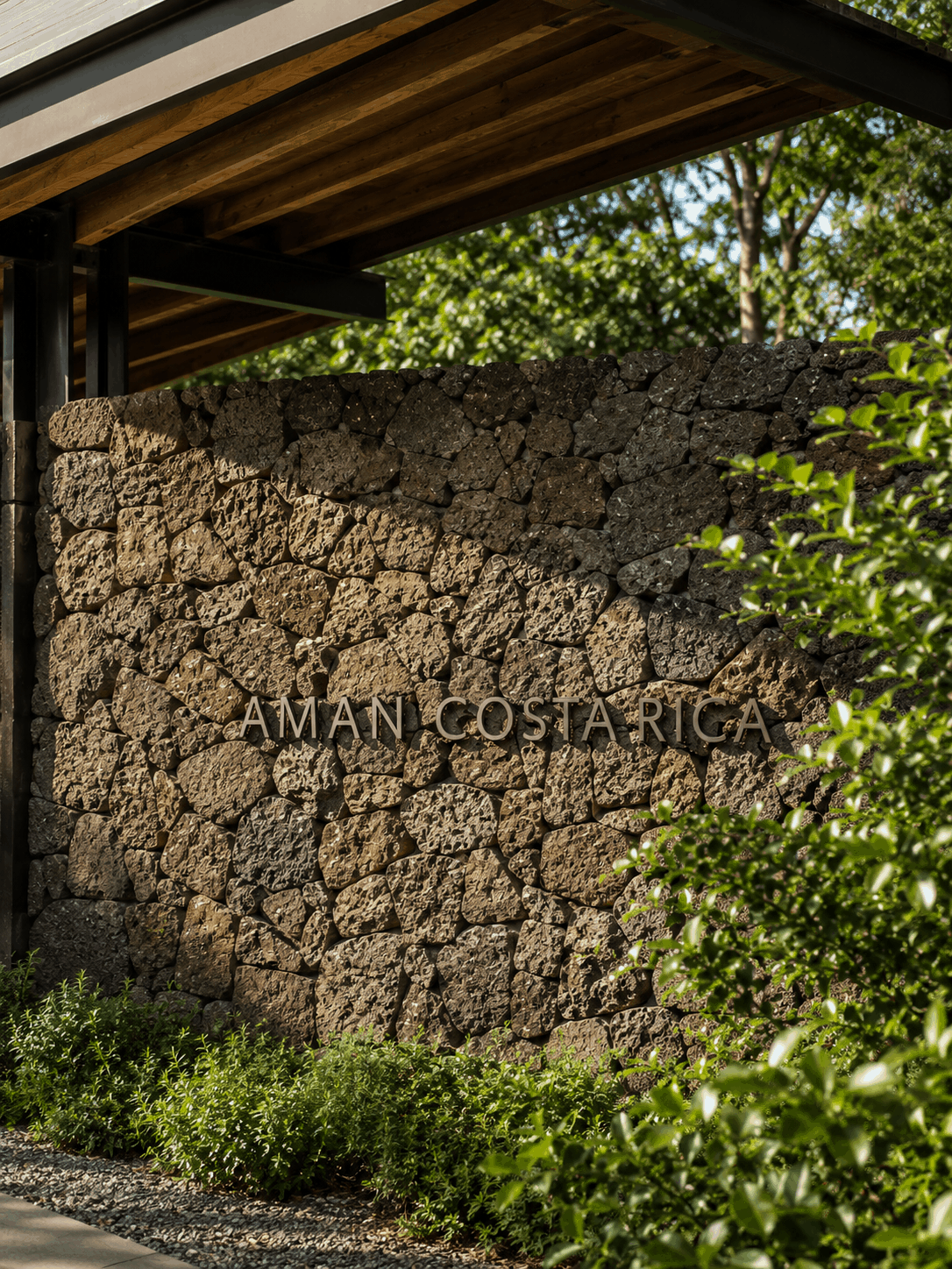 Close-up of 'Aman Costa Rica' lettering mounted on a rough stone wall, partially covered by vines and greenery.