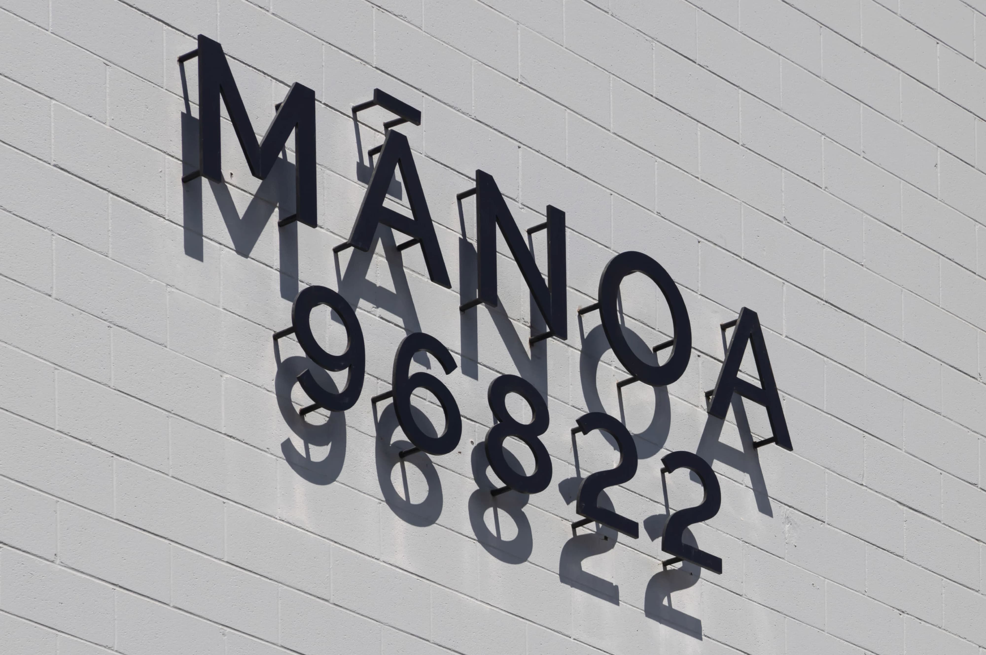 Dimensional black letters spelling 'Mānoa 96822' mounted diagonally on a white brick wall, casting long shadows in the sunlight.