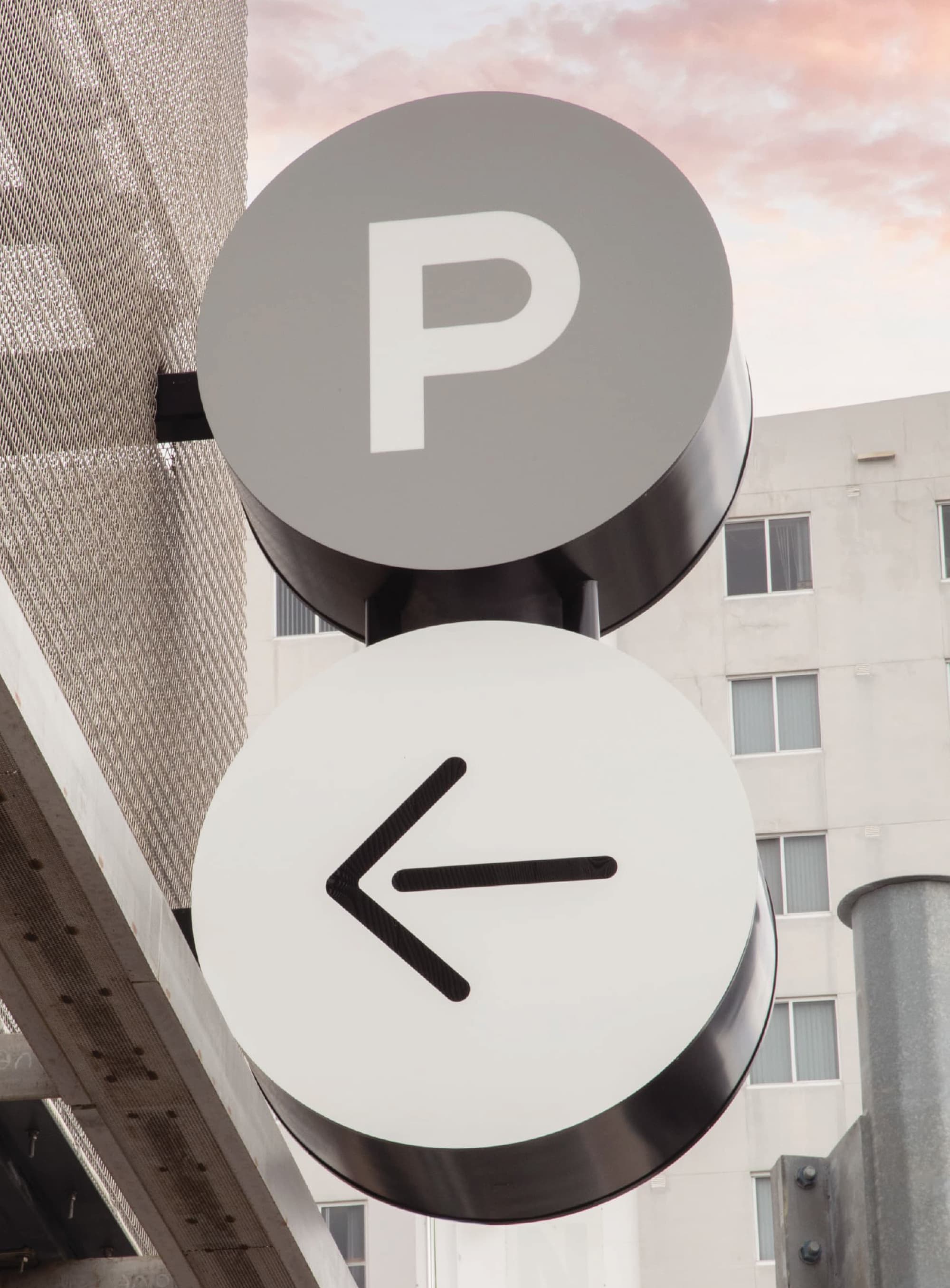Two round wayfinding signs mounted on a building exterior, the top gray disc displaying a white 'P' for parking and the bottom white disc displaying a black left-pointing arrow, with a residential building visible in the background at dusk.