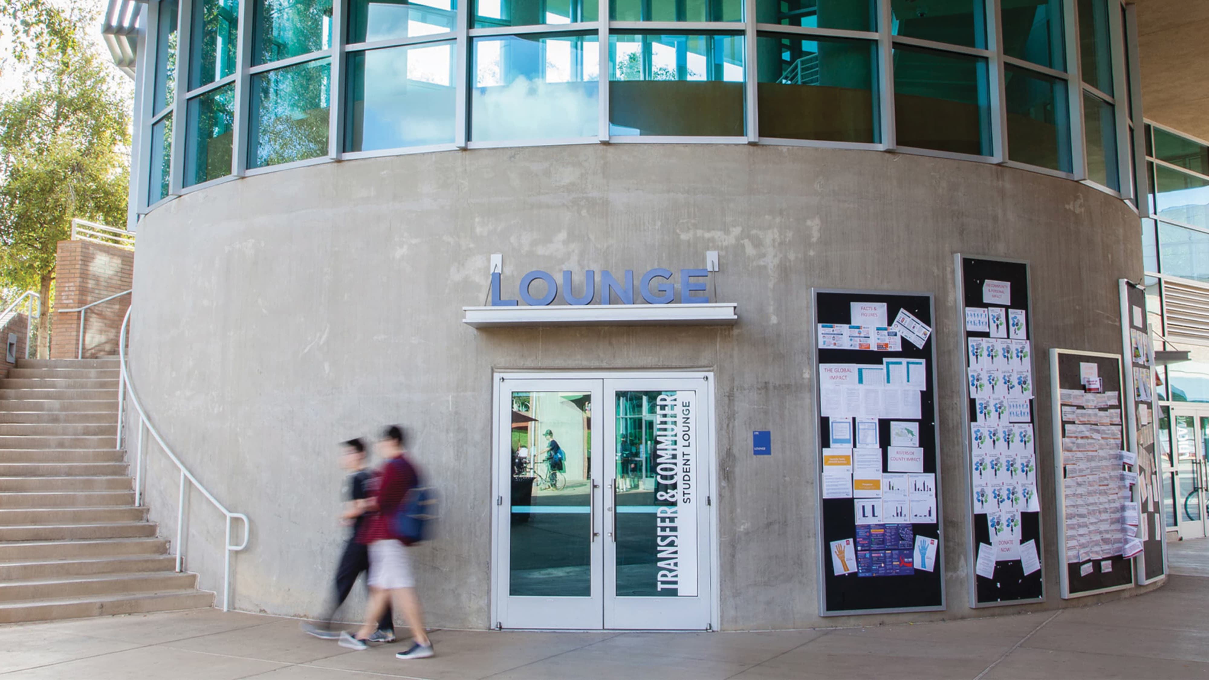 Curved concrete building exterior with a 'Lounge' sign above glass doors labeled 'Transfer & Commuter Student Lounge,' bulletin boards covered in flyers to the right, and two students walking past on the sidewalk.