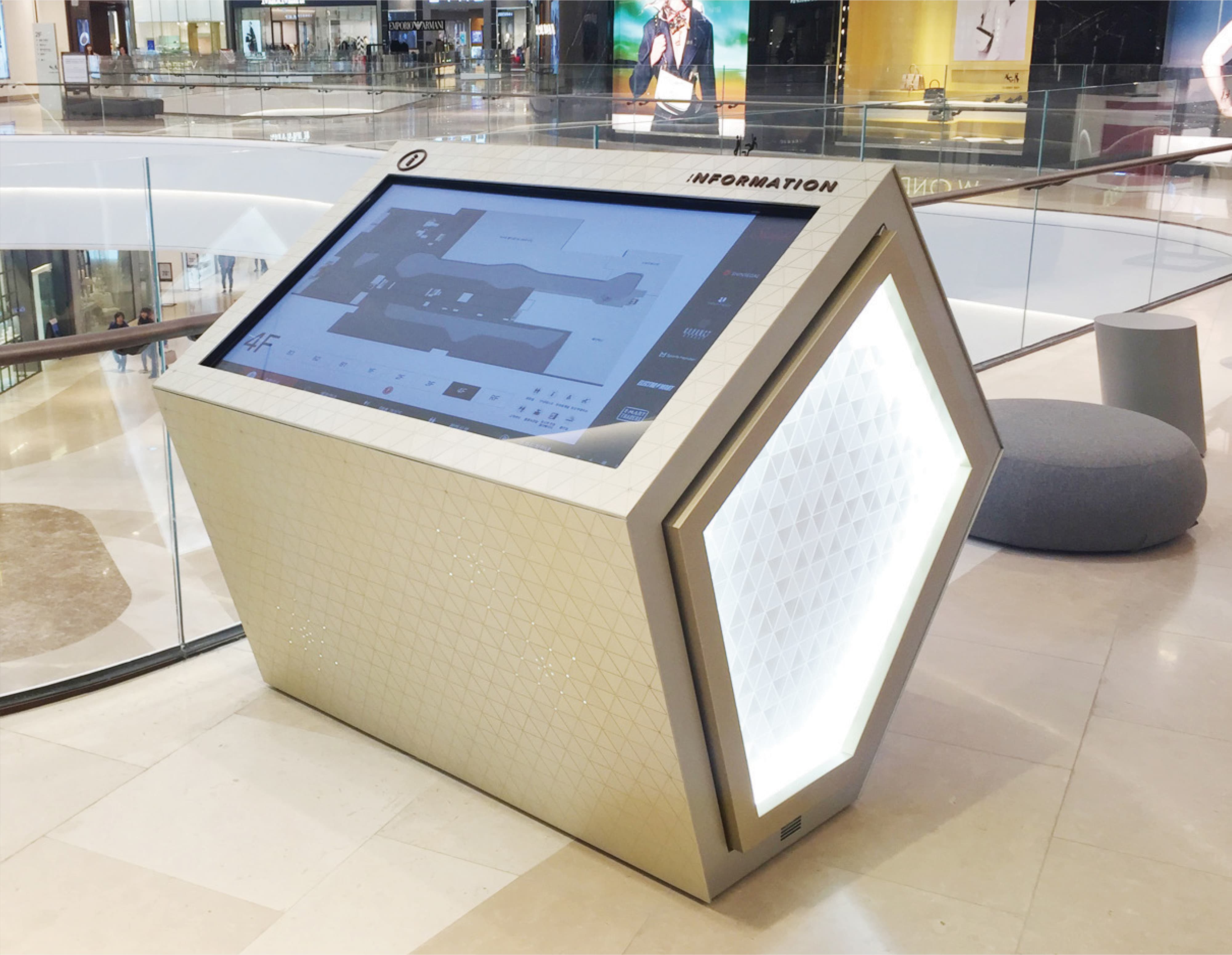Angled digital information kiosk with a touchscreen mall directory displaying a 4th floor map, a triangular gold-patterned housing, and an illuminated pentagon side panel, positioned in a mall atrium near storefronts and a curved glass railing
