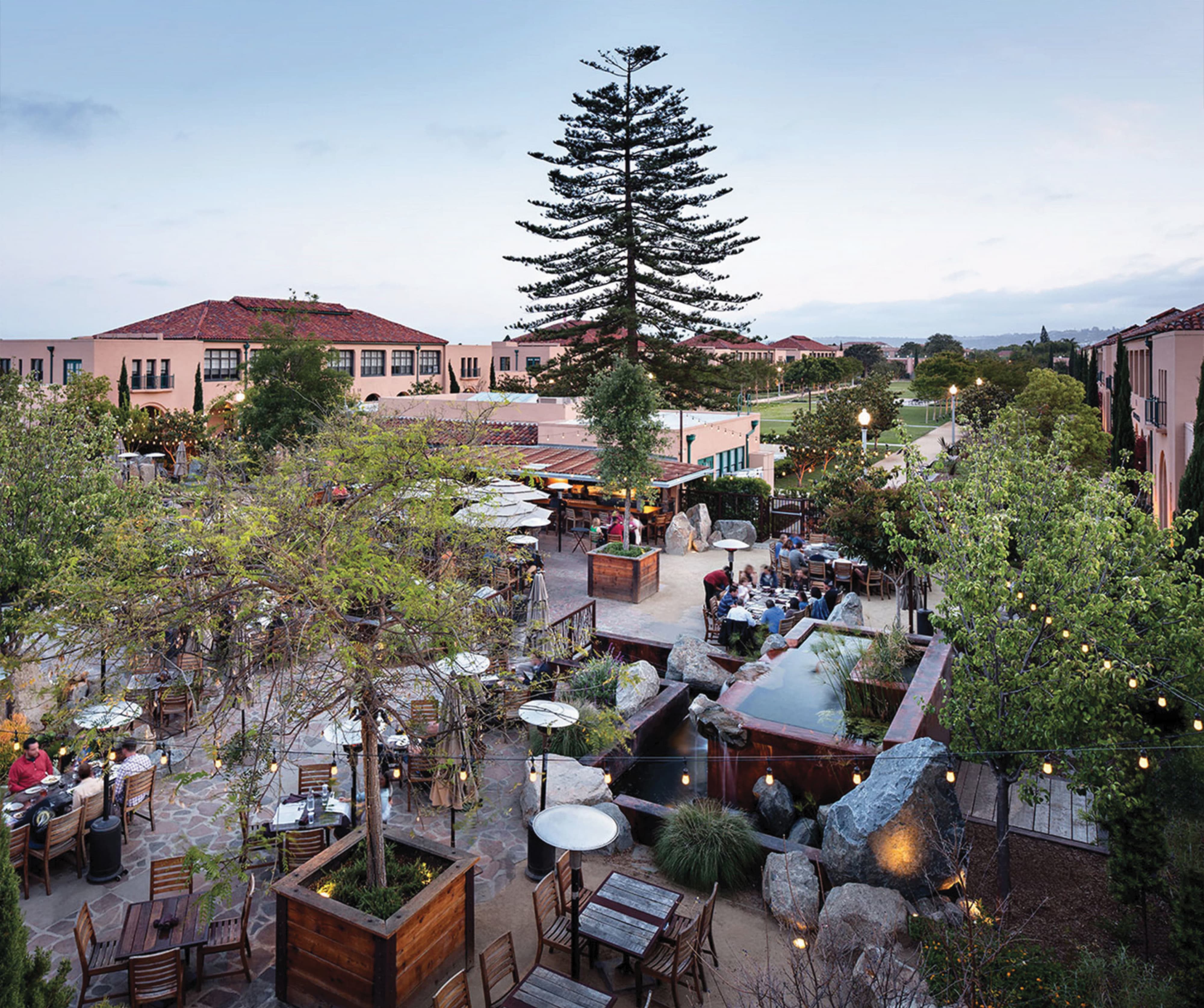 Elevated view of an outdoor dining patio at dusk with string lights, stone tables, boulder-lined water features, and diners seated among trees, framed by Spanish Colonial buildings and a tree-lined lawn beyond.