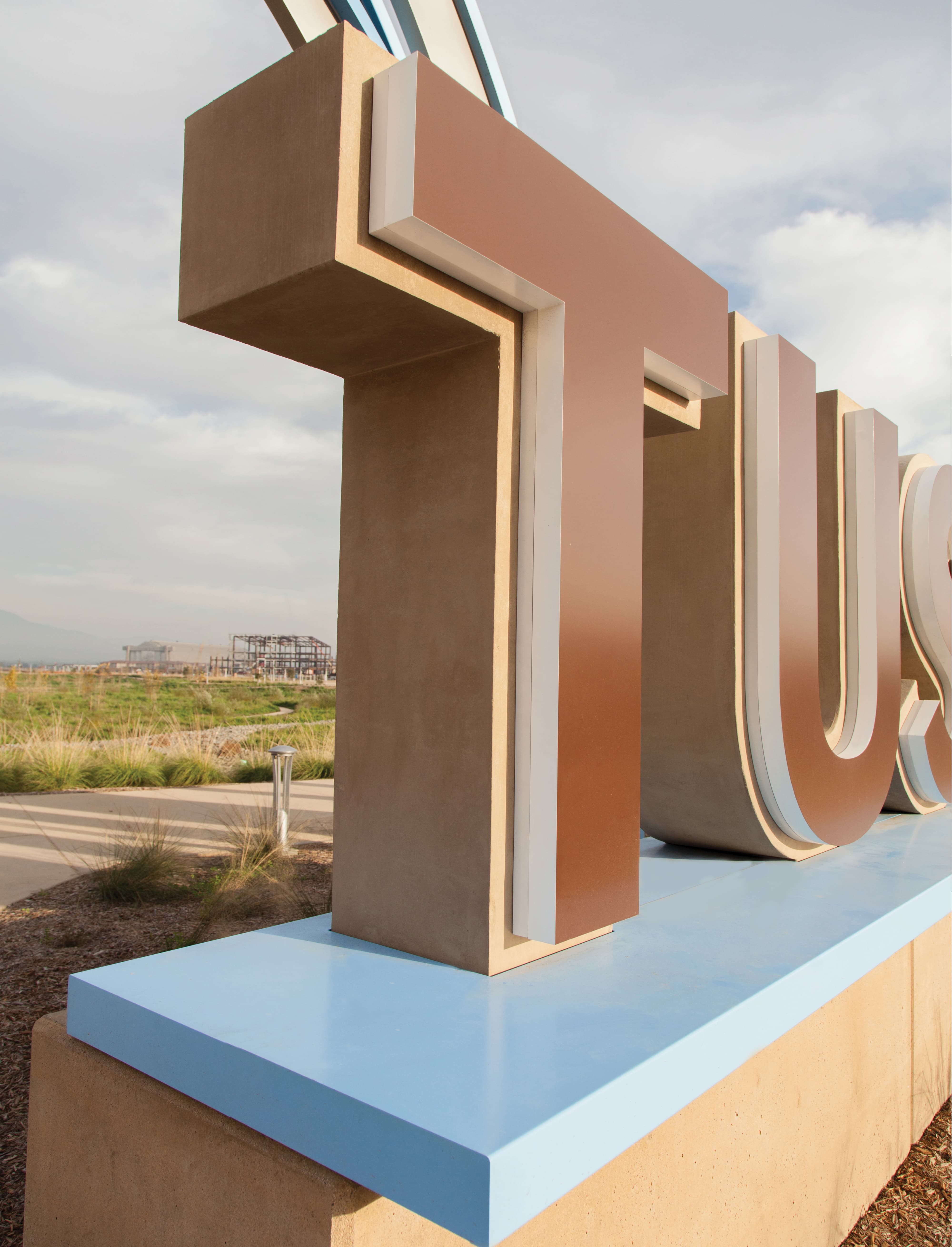 Close-up angled view of the 'T' and 'U' letters from a large dimensional 'Tustin' sign on a light blue base, with open grassland, a walking path, and a distant construction site visible in the background.
