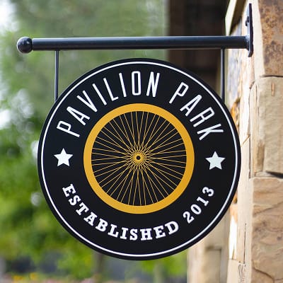 Circular blade sign for Pavilion Park featuring a gold bicycle wheel illustration and "Established 2013" text on a black background, wall-mounted on a stone exterior in Irvine, California, designed by RSM Design
