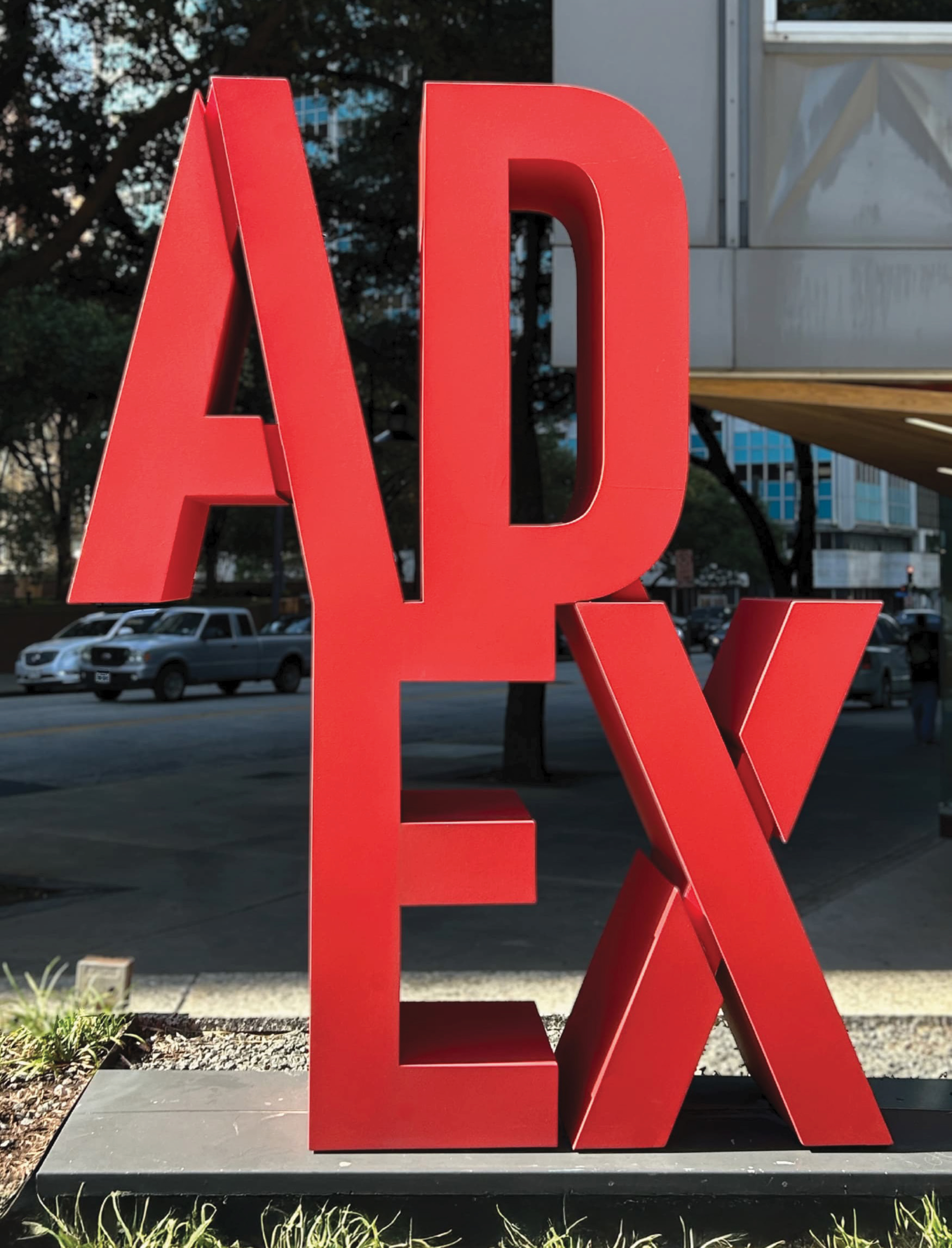 RSM Design large-scale exterior landmark identity sculpture for the Architecture and Design Exchange in downtown Dallas, featuring bold freestanding dimensional red metal ADEX letter forms positioned at the building entry alongside the wood canopy overhang and urban streetscape, demonstrating how environmental graphic design, public art, specialty graphics, and placemaking converge to create an iconic, human-centered landmark identity that commands attention, anchors a sense of place, and activates the pedestrian experience for cultural, civic, workplace, and streetscape markets.