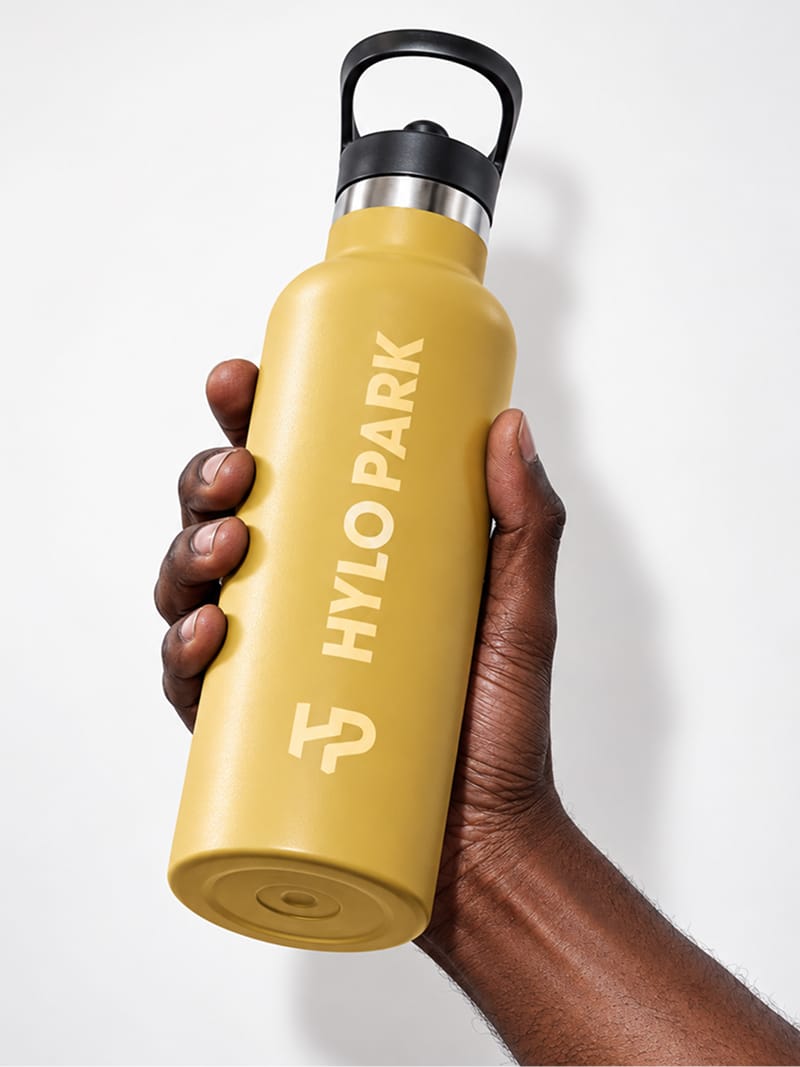 Hand holding a mustard-yellow insulated water bottle printed with the HP monogram and the HYLO PARK wordmark in cream, against a white background, Hylo Park, Las Vegas, brand identity design by RSM Design.