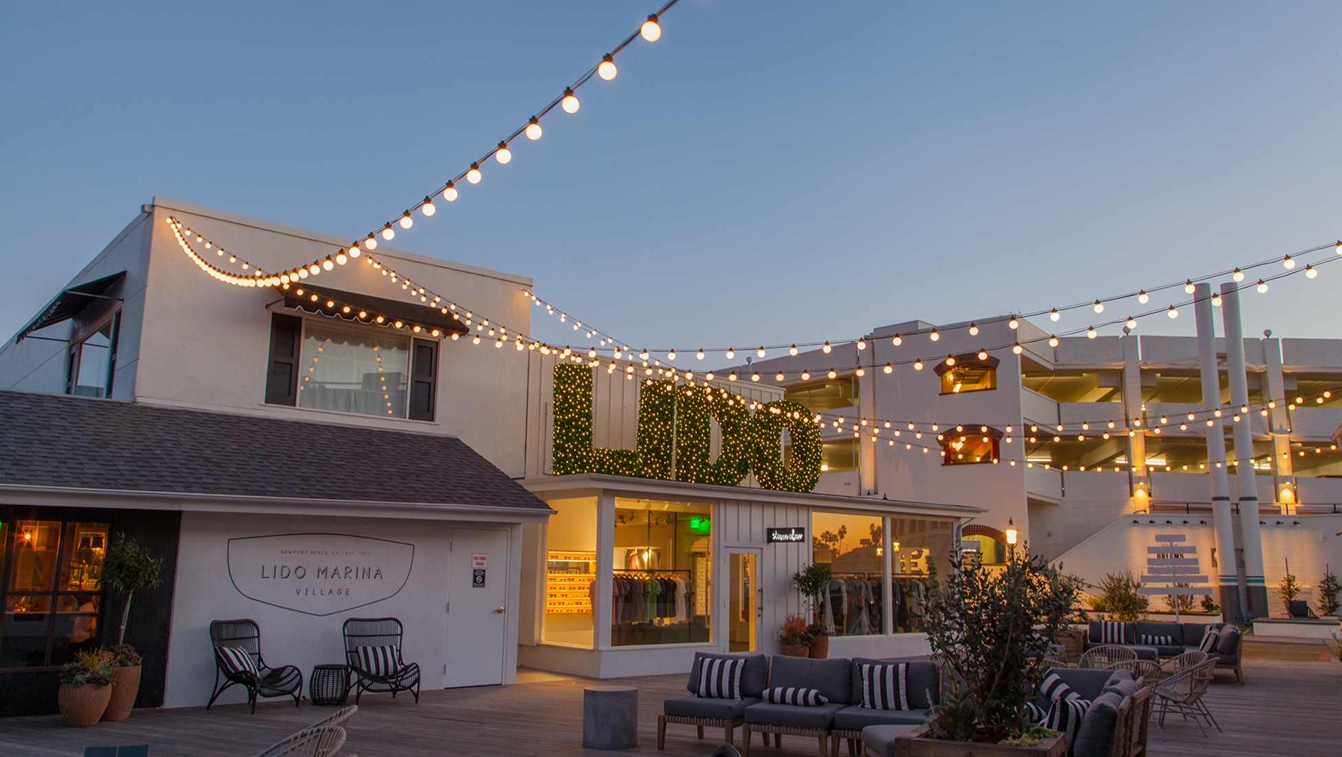 Lido Marina Village outdoor courtyard at dusk with string lights, illuminated boxwood "LIDO" topiary letters, waterfront boutique shops, and outdoor seating lounge area in Newport Beach, California