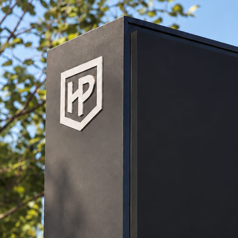 Close-up of a dimensional brushed metal HP shield mark mounted on the textured dark corner of a sign, set against trees and blue sky, Hylo Park, Las Vegas, wayfinding design by RSM Design.