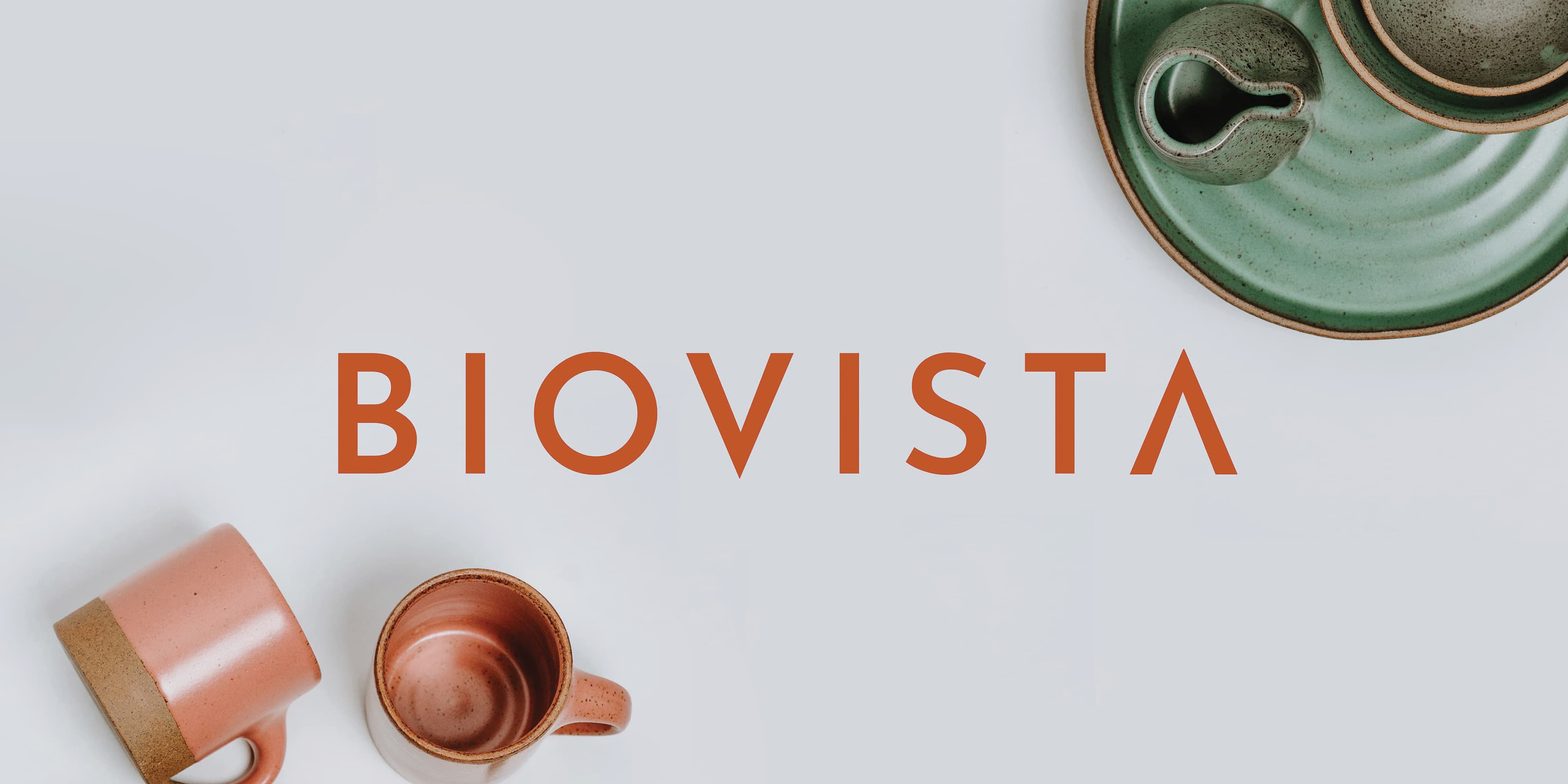 Orange 'Biovista' wordmark centered on a light gray background, surrounded by ceramic pottery including two terracotta mugs and a green plate with a small pitcher.