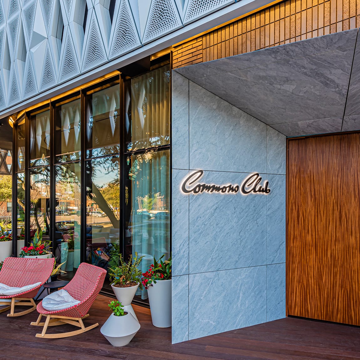 Commons Club restaurant entrance at Virgin Hotels Dallas featuring backlit cursive channel letter signage on marble facade with wood door and outdoor patio seating - interior and exterior hospitality signage and environmental branding design by RSM Design, a leading wayfinding and experiential graphics firm
